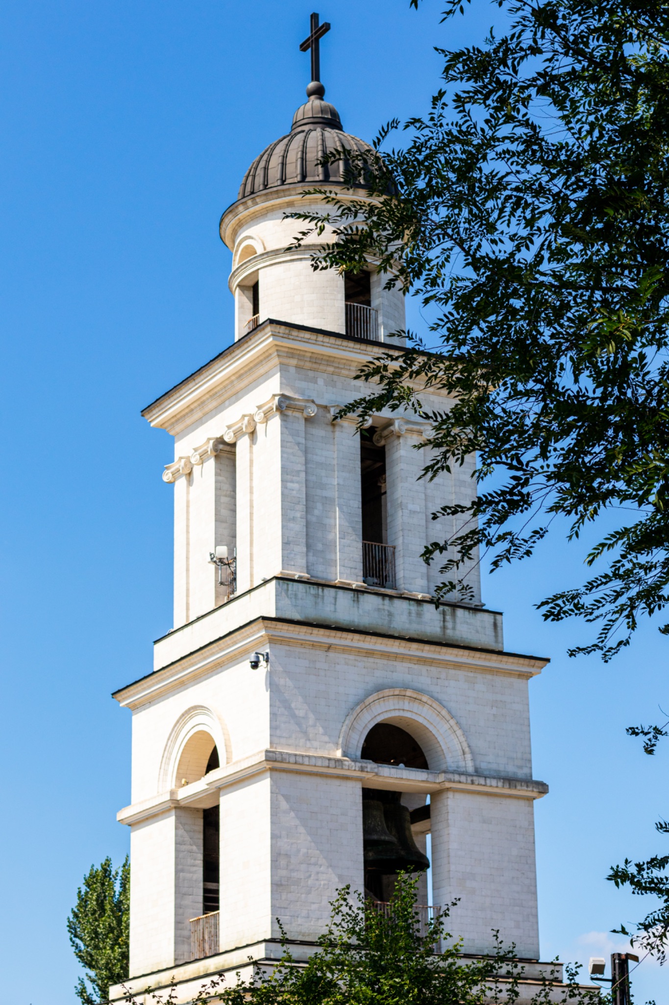 White church bell tower with a dome and cross against a clear blue sky, framed by green foliage in Chisinau, Moldova.