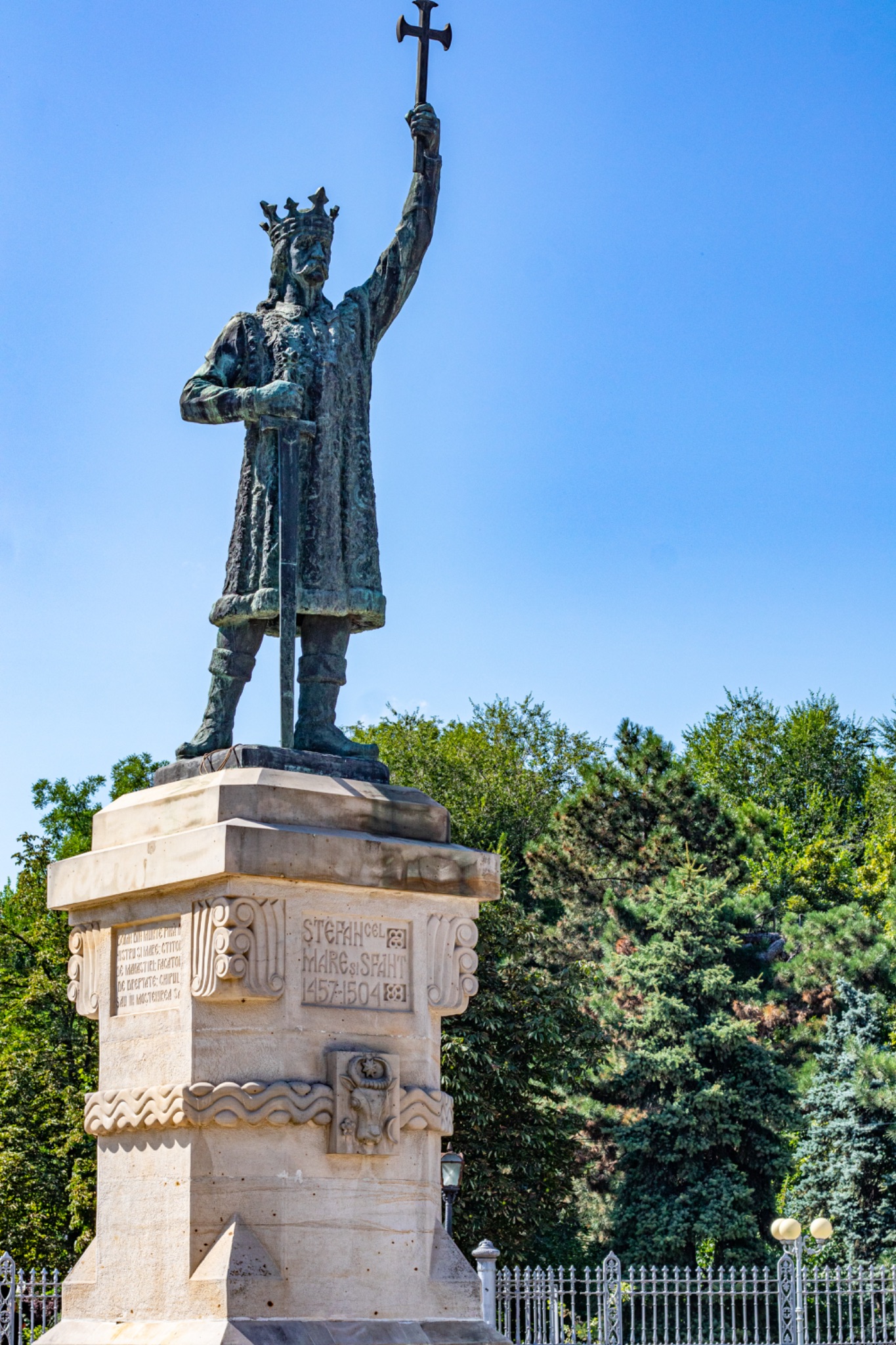 Stefan Cel Mare statue of a historical figure with a cross, set against a clear blue sky and lush greenery in Chisinau, Moldova.