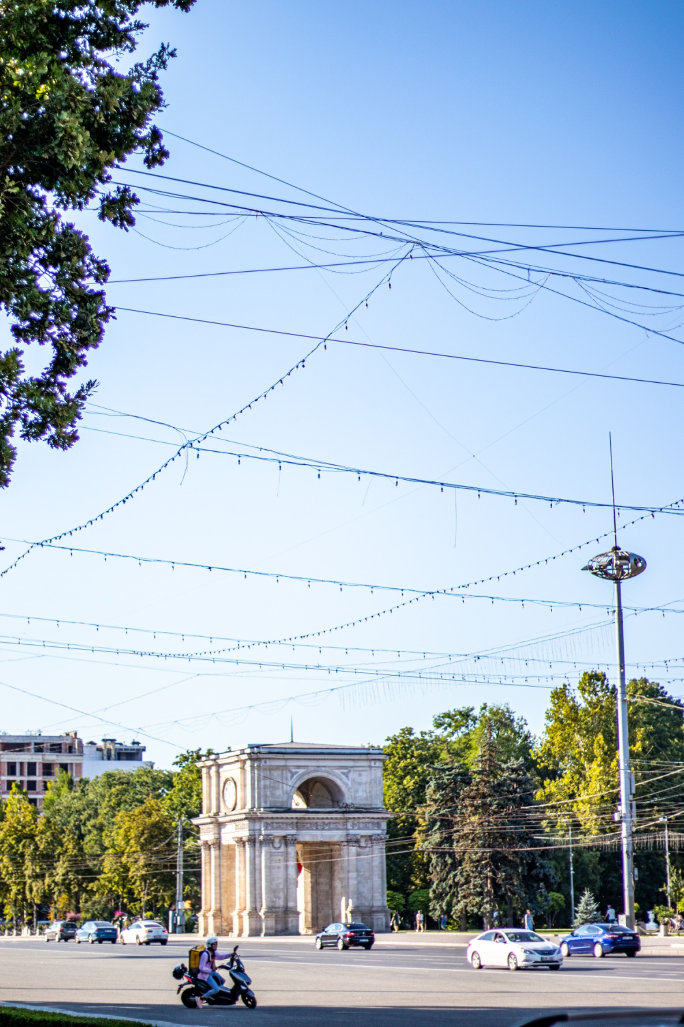 Urban street scene with power lines, a historical arch monument in the background, and vehicles in motion under a clear blue sky in Chisinau, Moldova.
