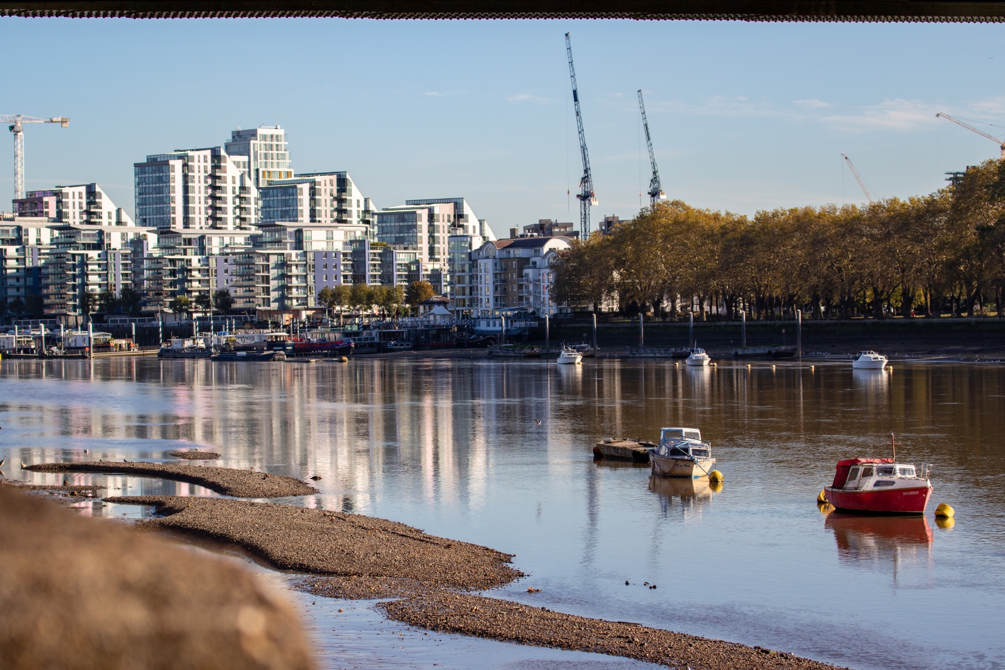 Tranquil river scene with moored boats and modern city skyline in the background on a sunny day.