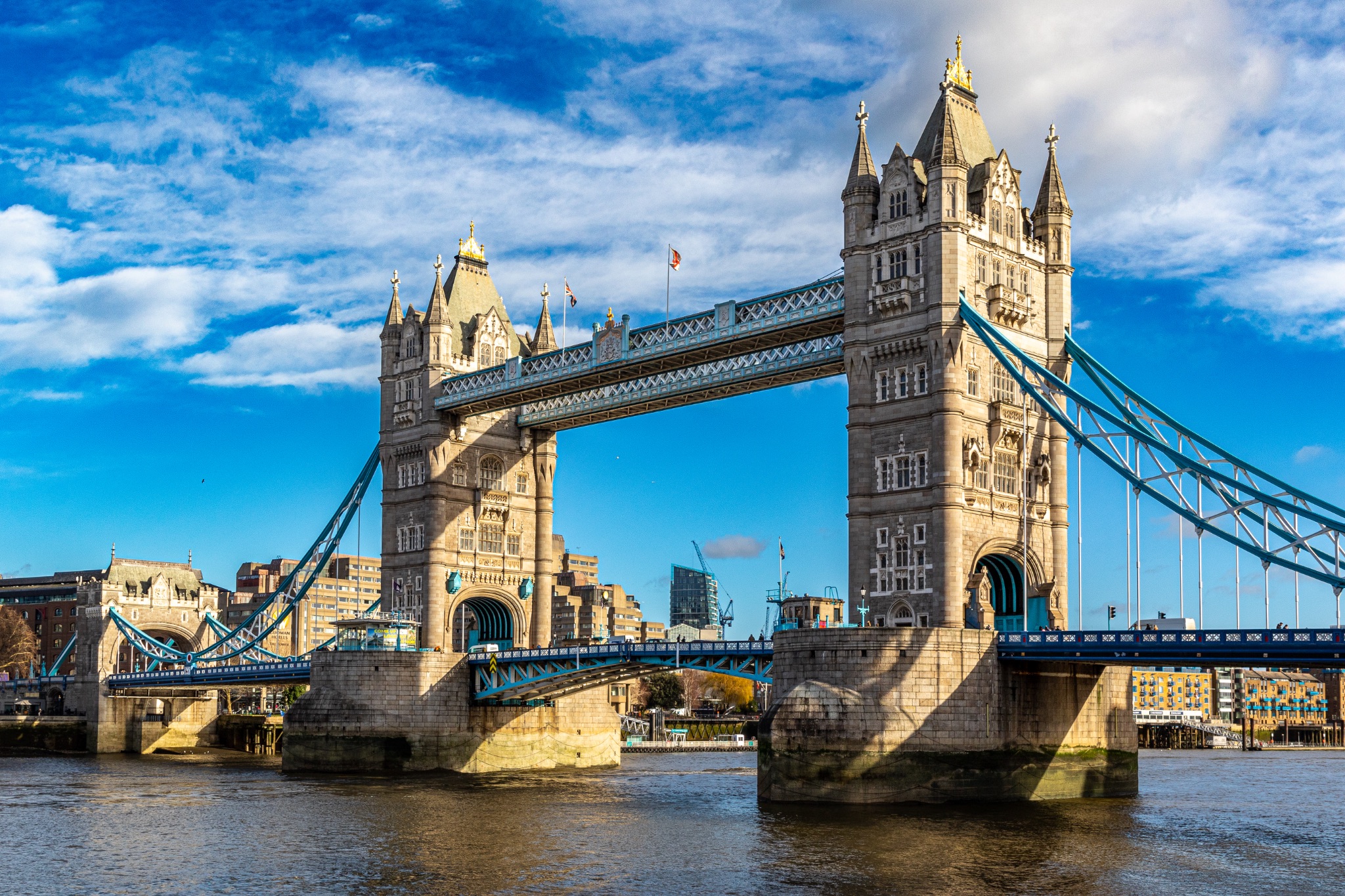 Sunny day view of the iconic Tower Bridge over the River Thames in London, UK, with blue skies and fluffy clouds.