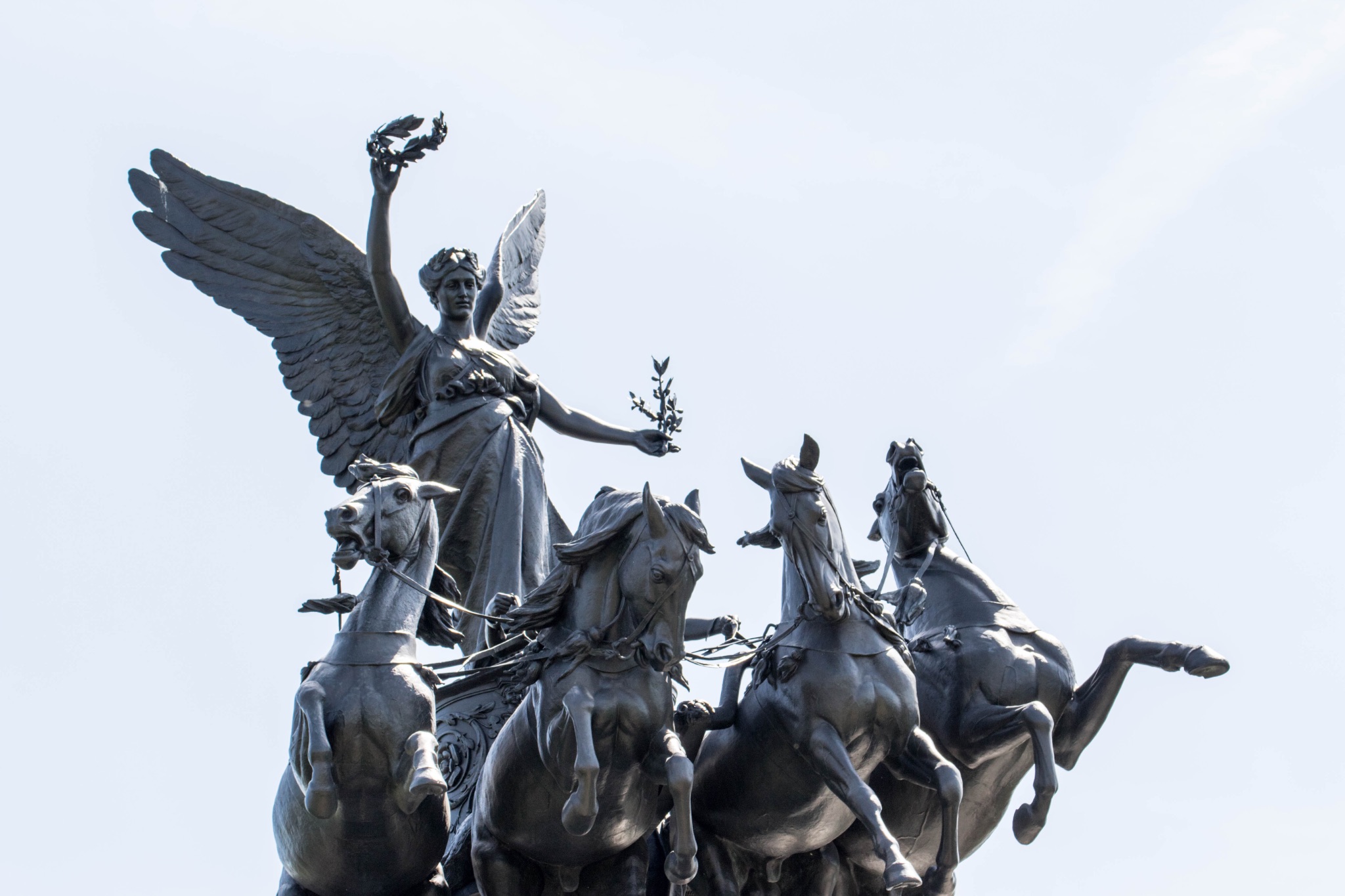 Bronze sculpture of a winged female figure with a laurel wreath, leading a chariot pulled by four horses against a clear sky.