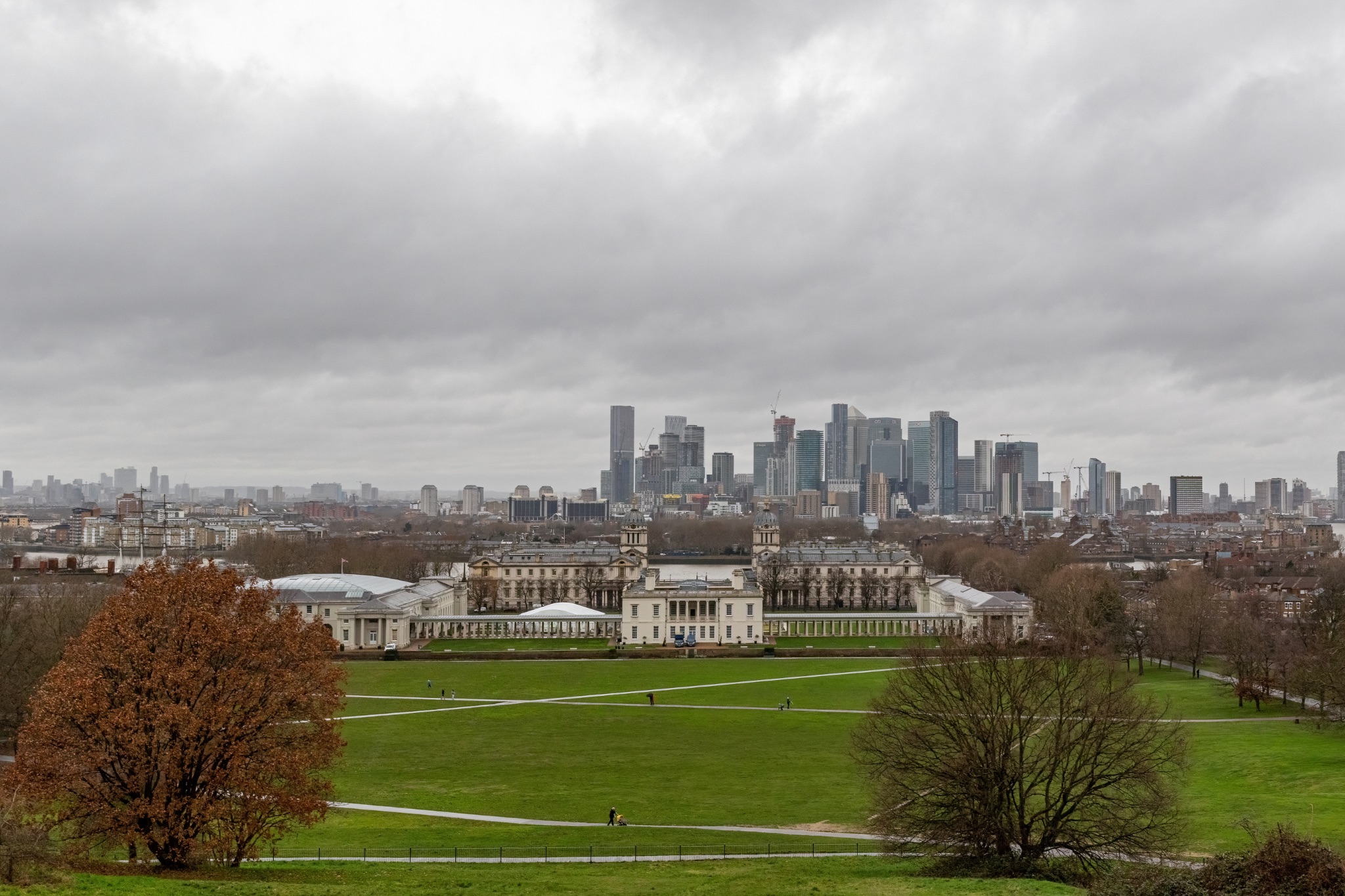 Panoramic view of a city skyline with modern skyscrapers behind a large green park under a cloudy sky.