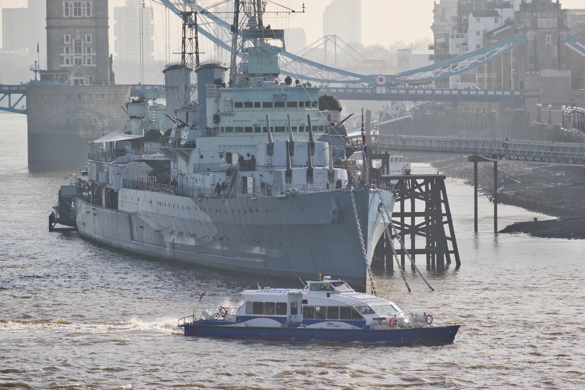 Misty view of a warship moored by a city river with a small boat passing by.