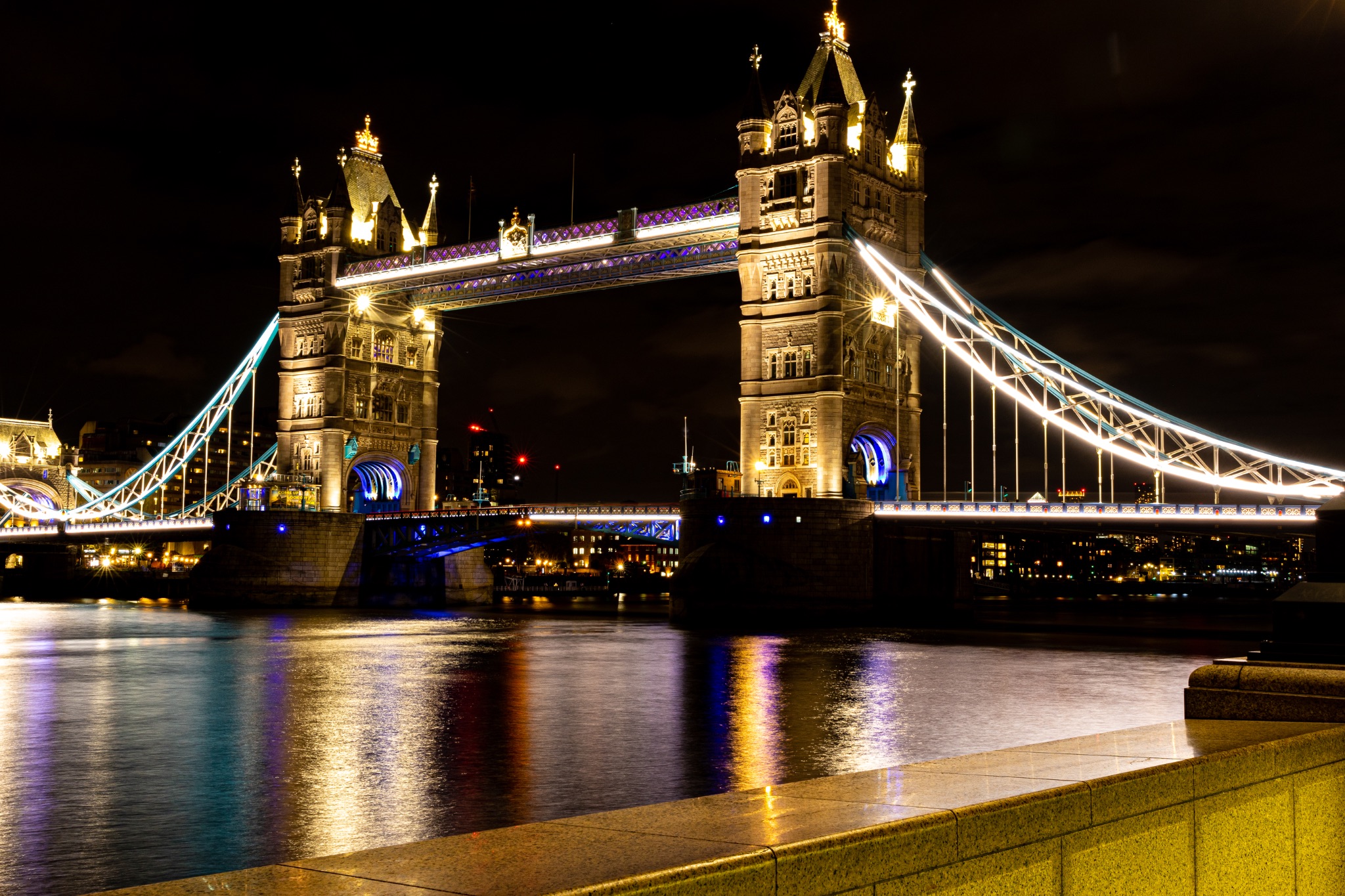 Night view of the illuminated Tower Bridge in London over the Thames River with reflections on water.