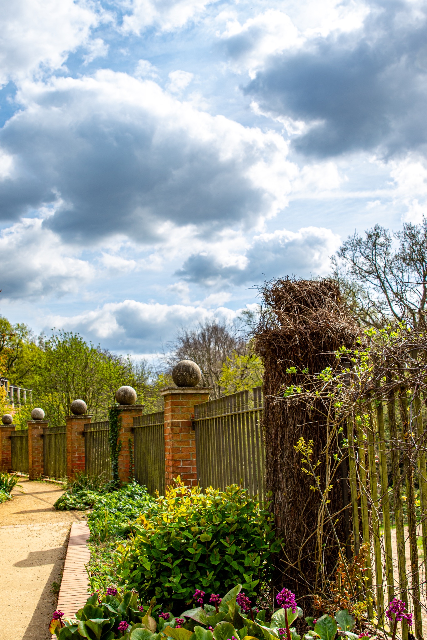 Pathway in a serene garden with blooming flowers, a wooden fence, and a cloudy blue sky in The Hill Garden and Pergola in London.