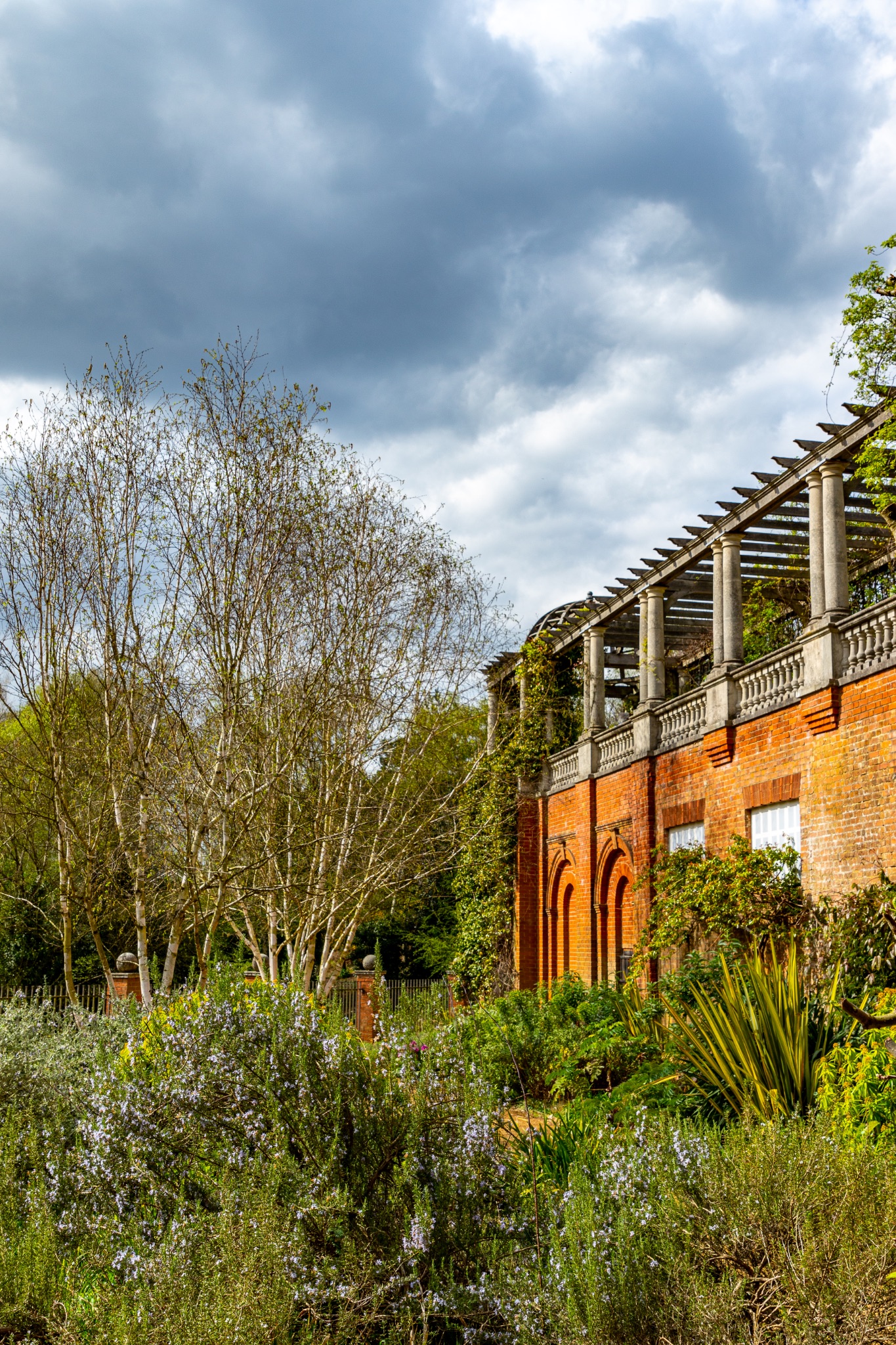 Scenic view of a lush garden with diverse flora in front of a rustic building with a pergola, under a cloudy sky in The Hill Garden and Pergola in London.