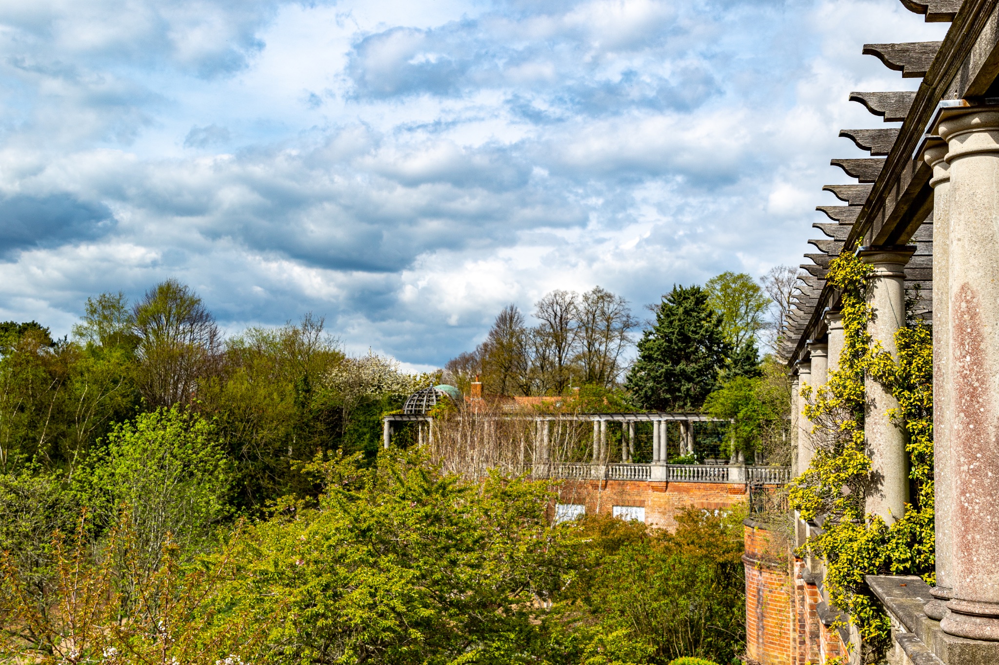 Scenic view of a lush garden with classical architecture under a partly cloudy sky in The Hill Garden and Pergola in London.
