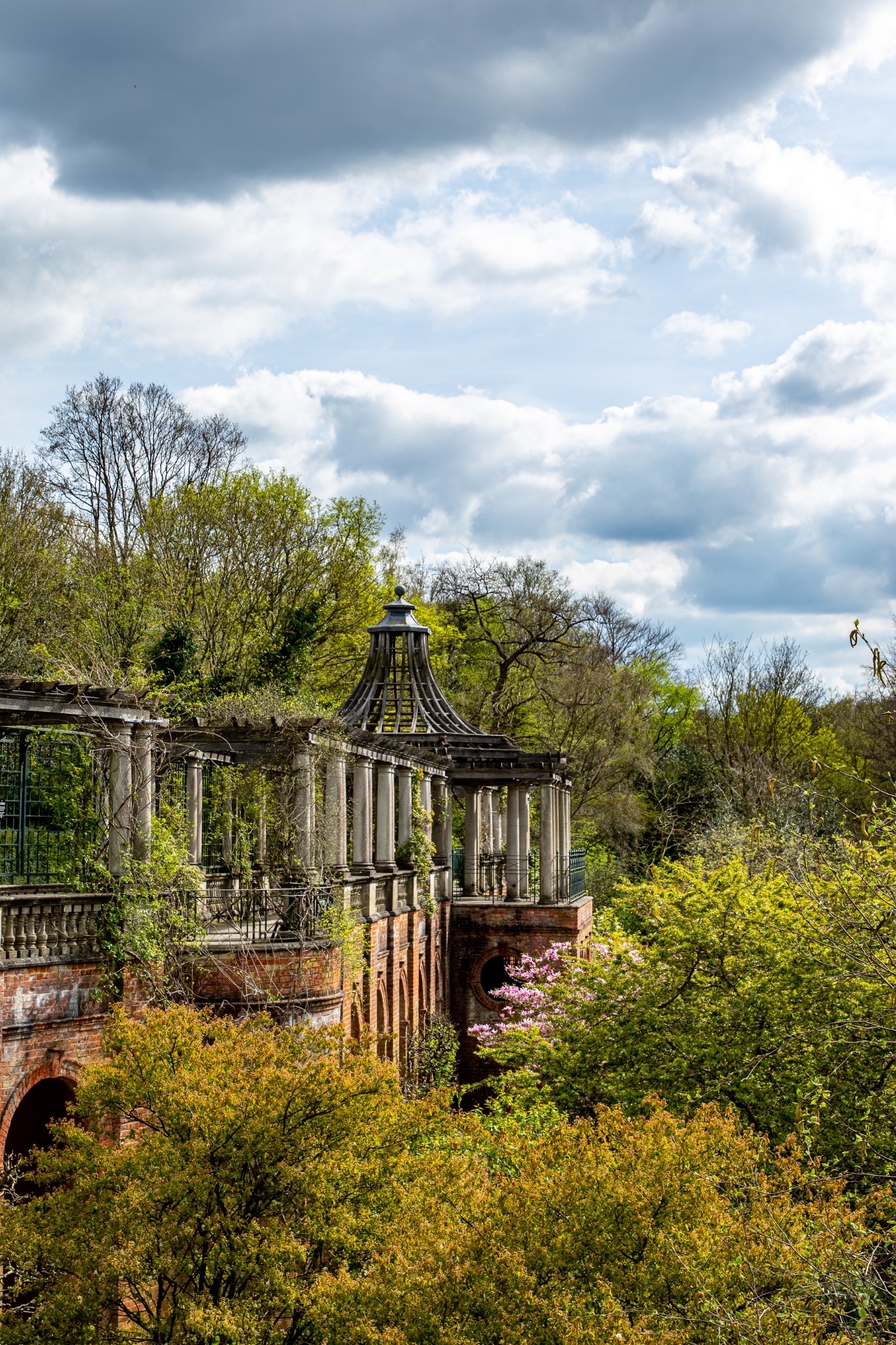 Ruined classical architecture with columns amidst lush greenery under a cloudy sky in The Hill Garden and Pergola in London.