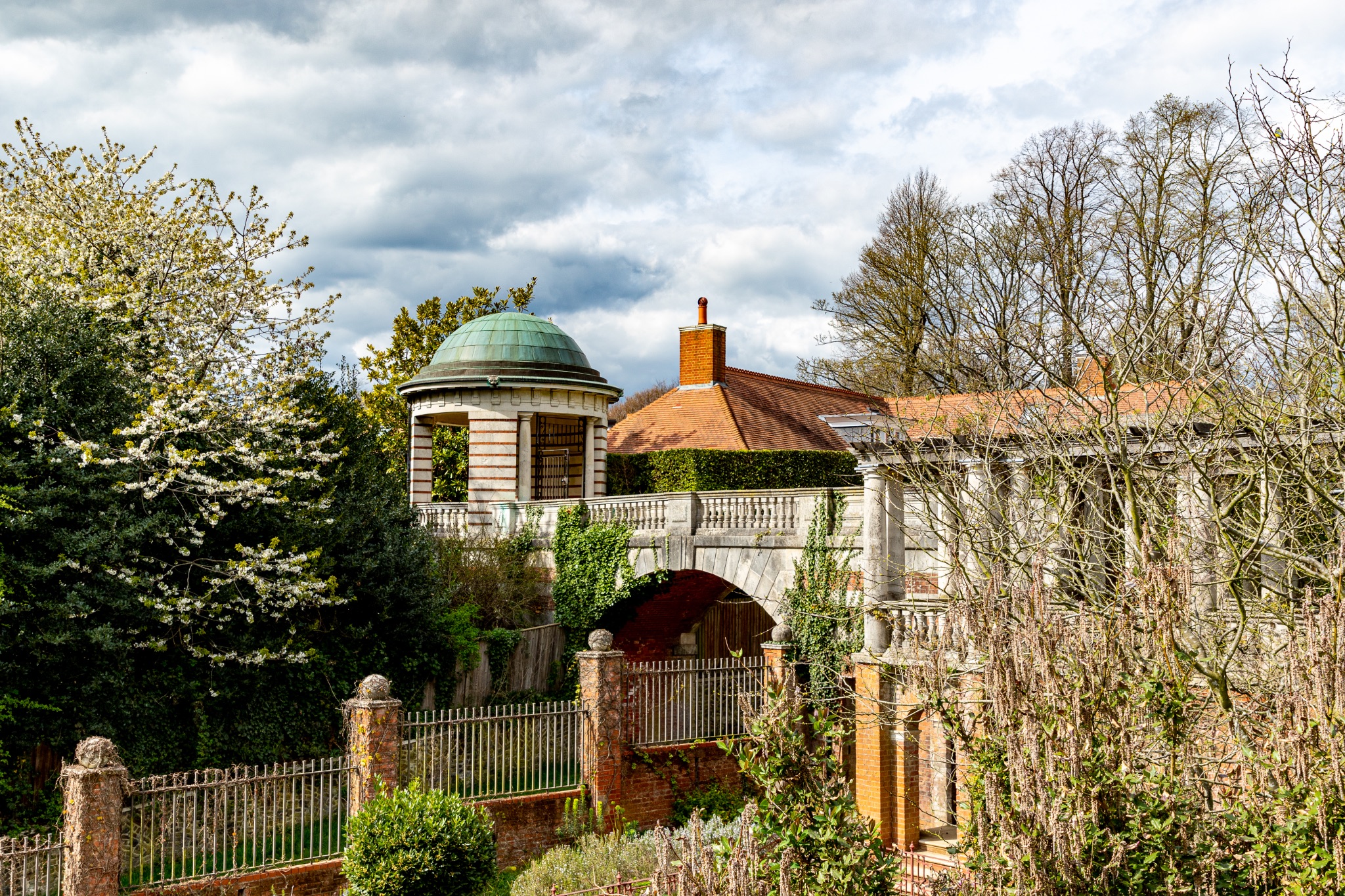 Quaint old house with a dome and archway surrounded by lush greenery under a cloudy sky in The Hill Garden and Pergola in London.