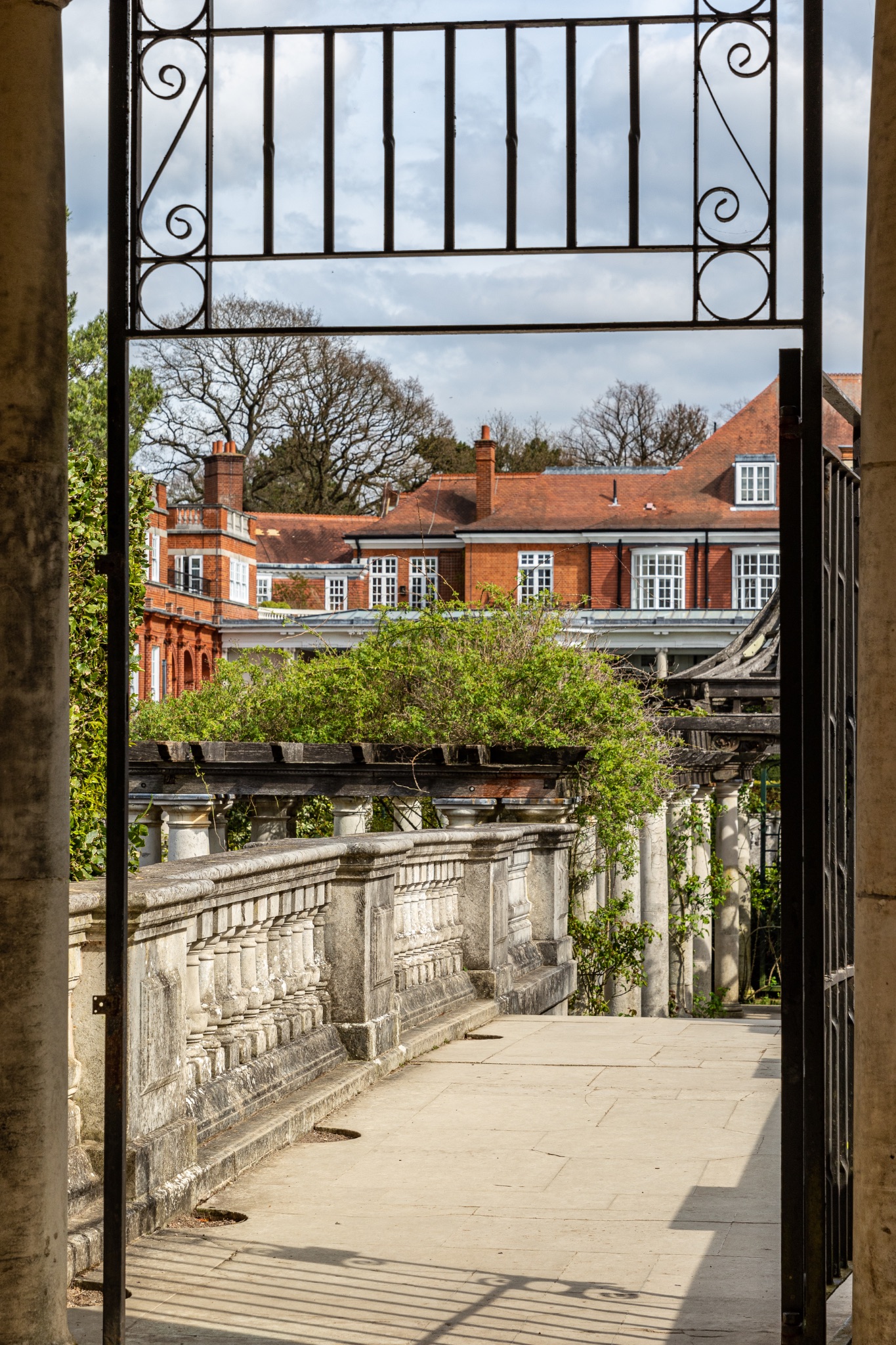 Elegant estate viewed through an open gate, showcasing lush greenery and classic architecture under a blue sky with clouds in The Hill Garden and Pergola in London.