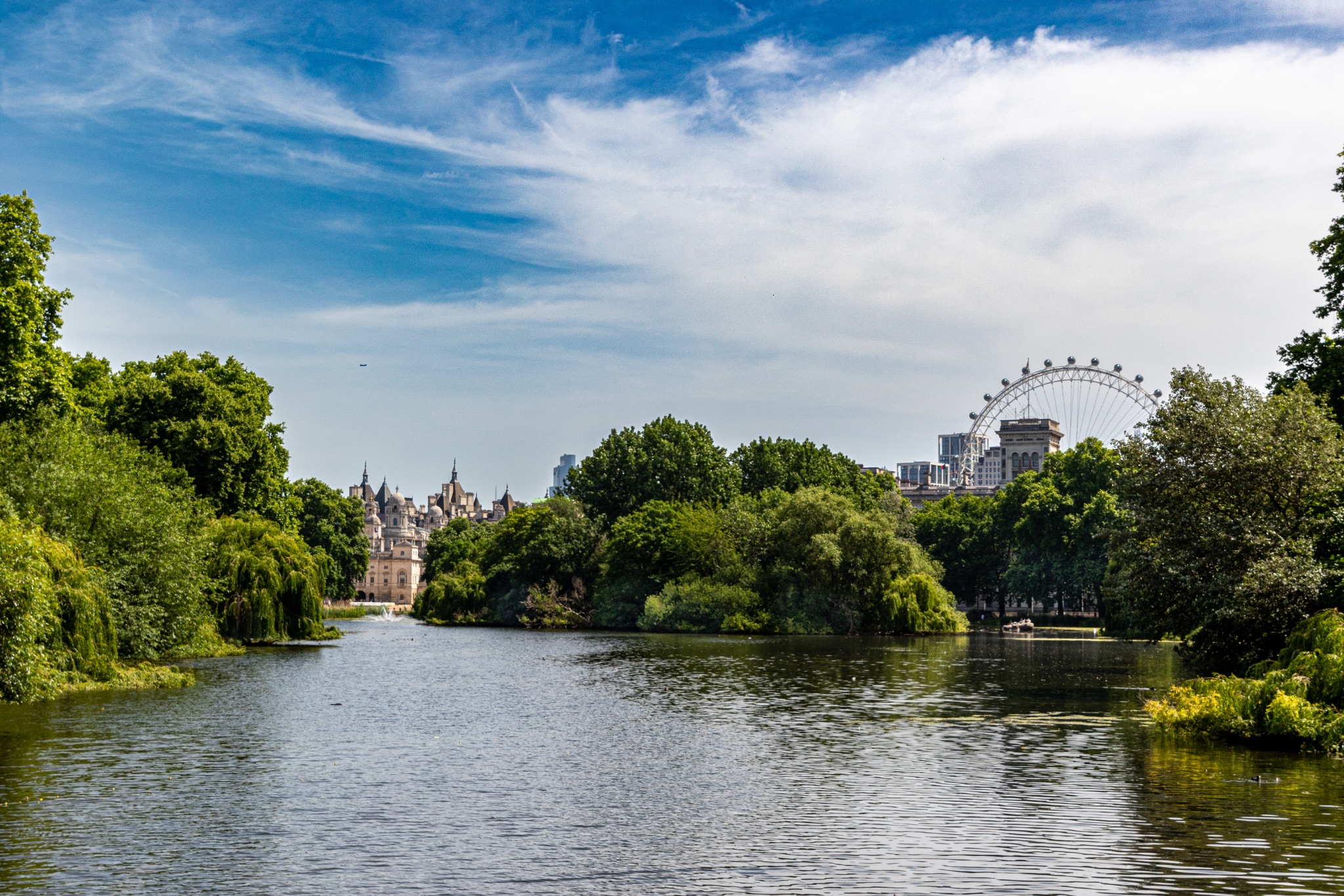 Scenic view of a tranquil river with lush greenery and a city skyline featuring a large Ferris wheel in the background.