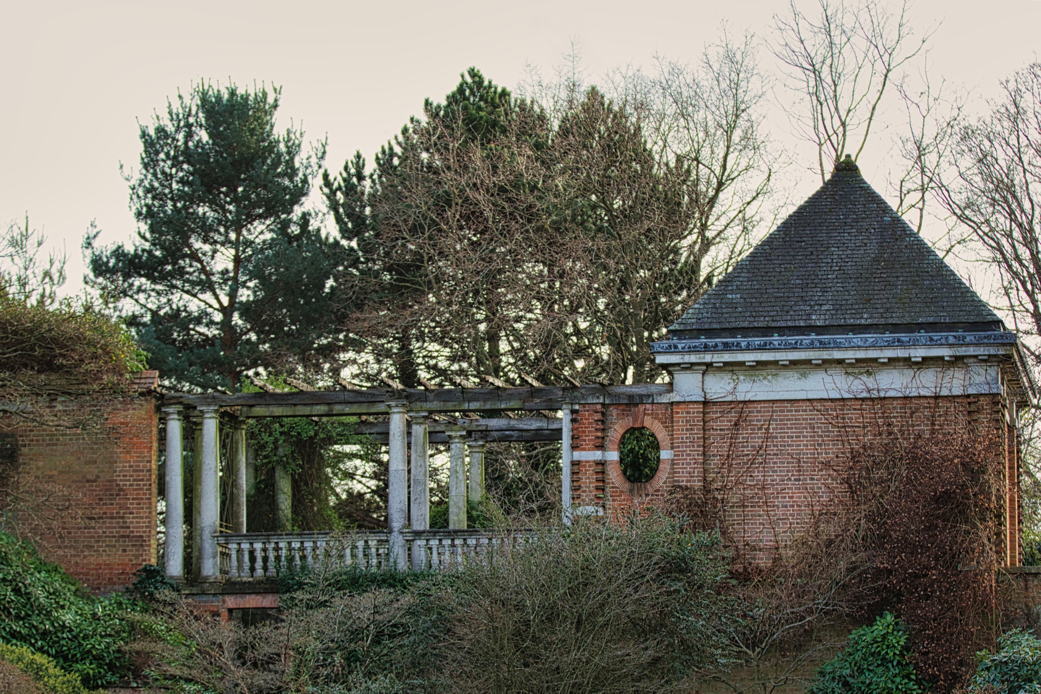 Abandoned brick building with overgrown vegetation and a classic columned structure, conveying a sense of neglect and history.