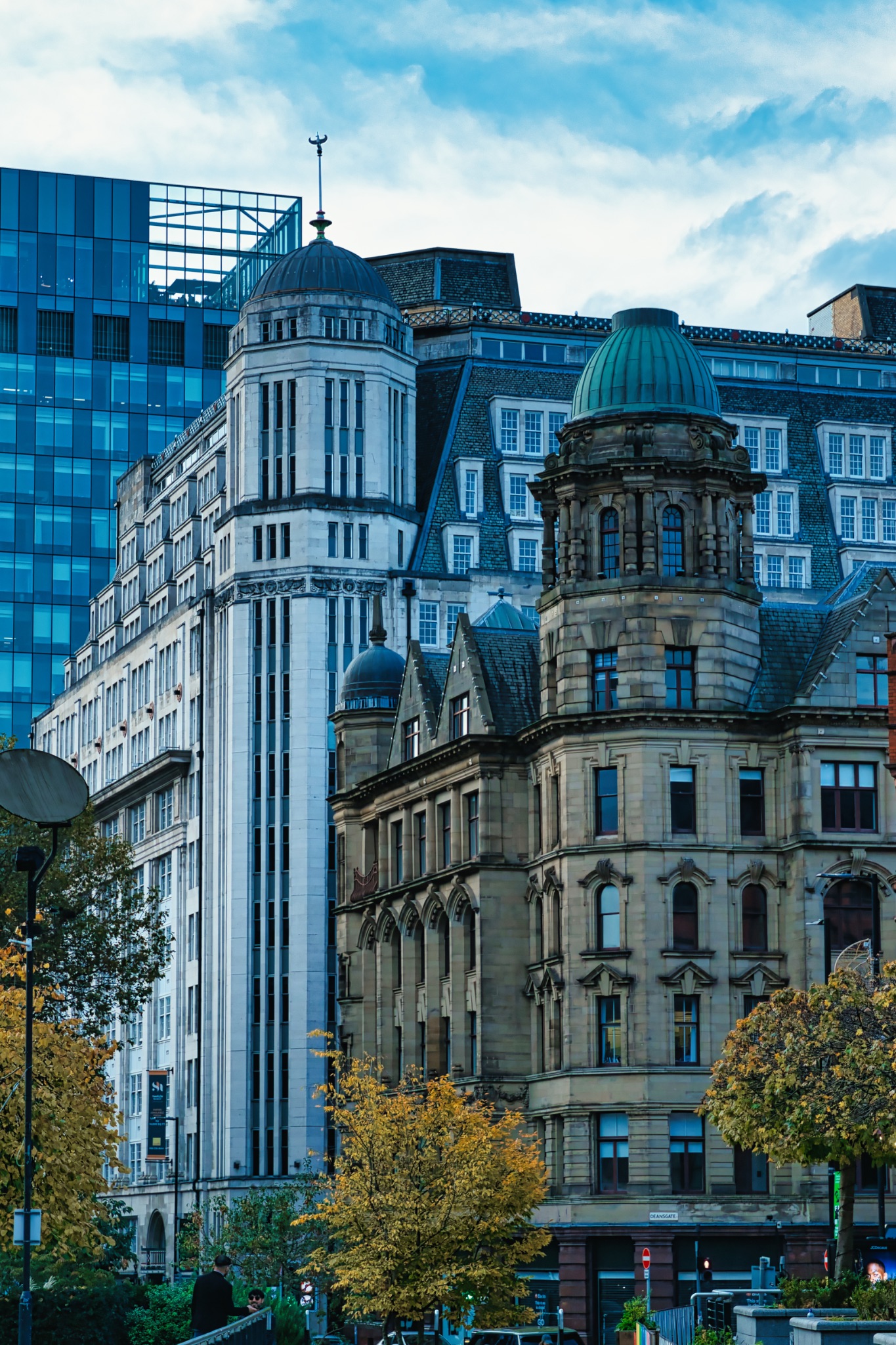 Contrast of architectural styles in a cityscape with modern and classical buildings against a blue sky with green foliage in the foreground in Manchester, England.