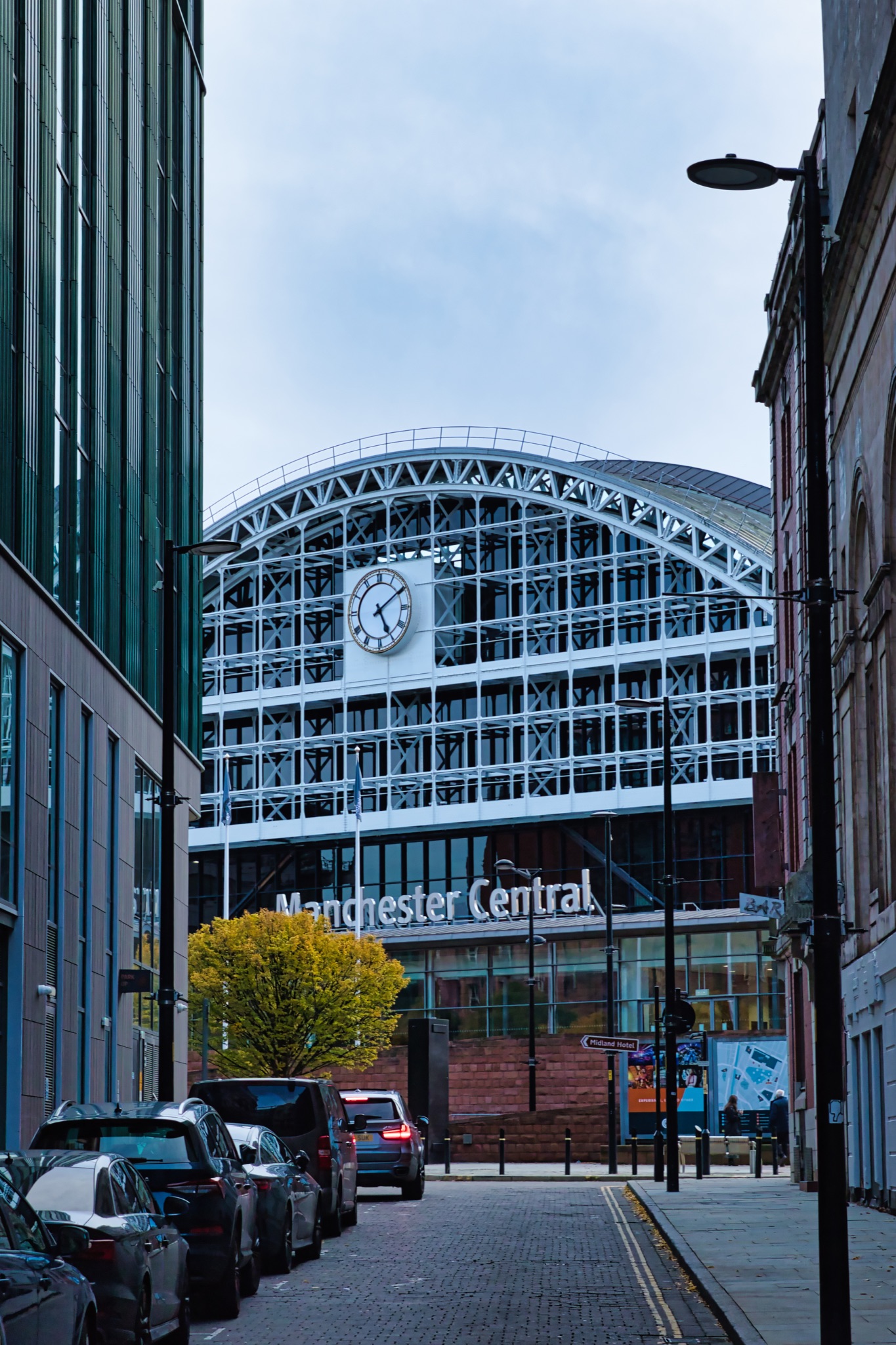 Modern architecture of Manchester Central Convention Complex with a prominent clock, viewed from a street lined with parked cars in Manchester, England.