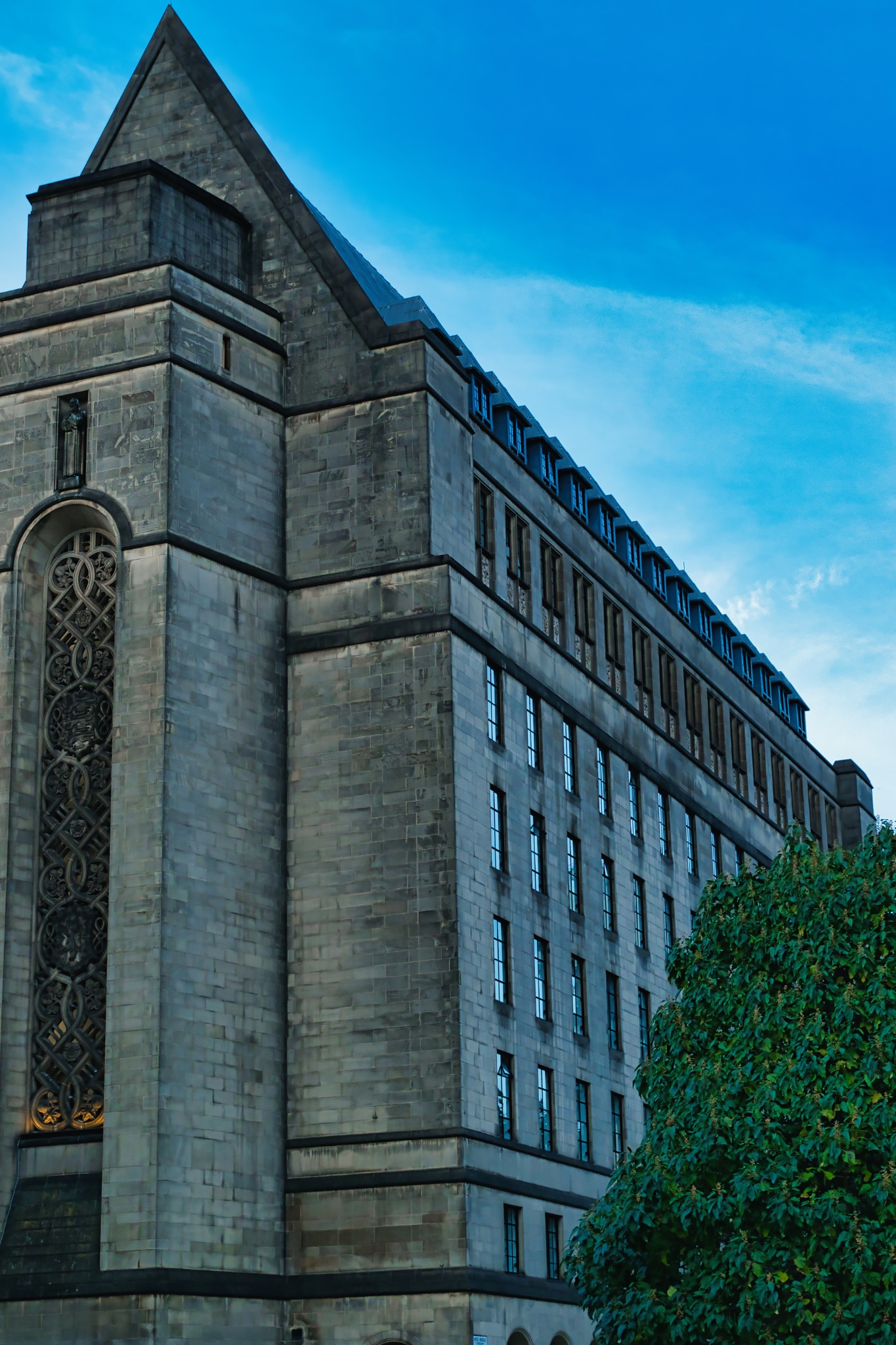 Architectural detail of a modern building with ornate windows against a clear blue sky, surrounded by green foliage in Manchester, England.