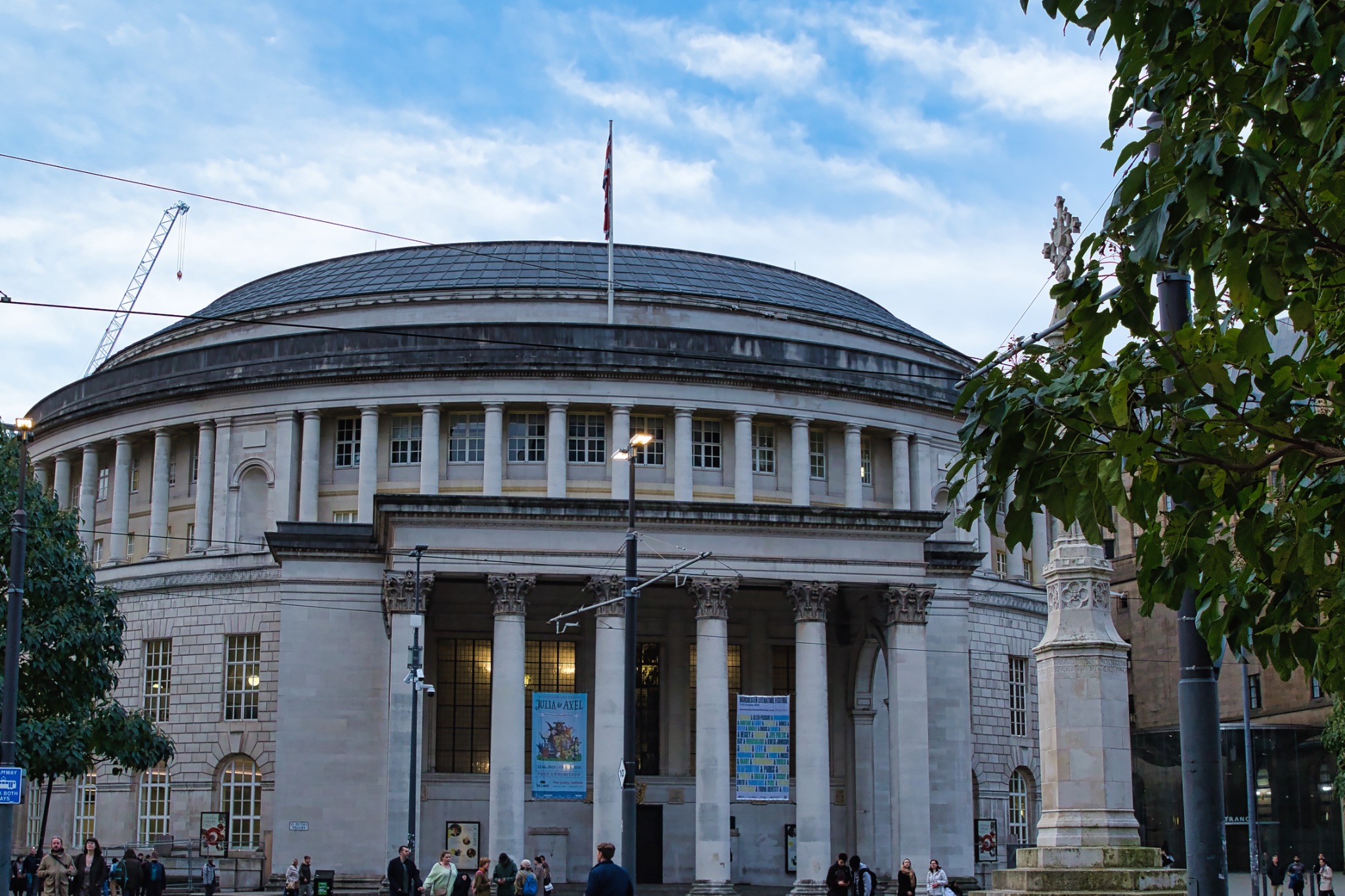 Exterior view of a classical building with columns and a dome, under a blue sky with clouds, people visible at the entrance in Manchester, England.