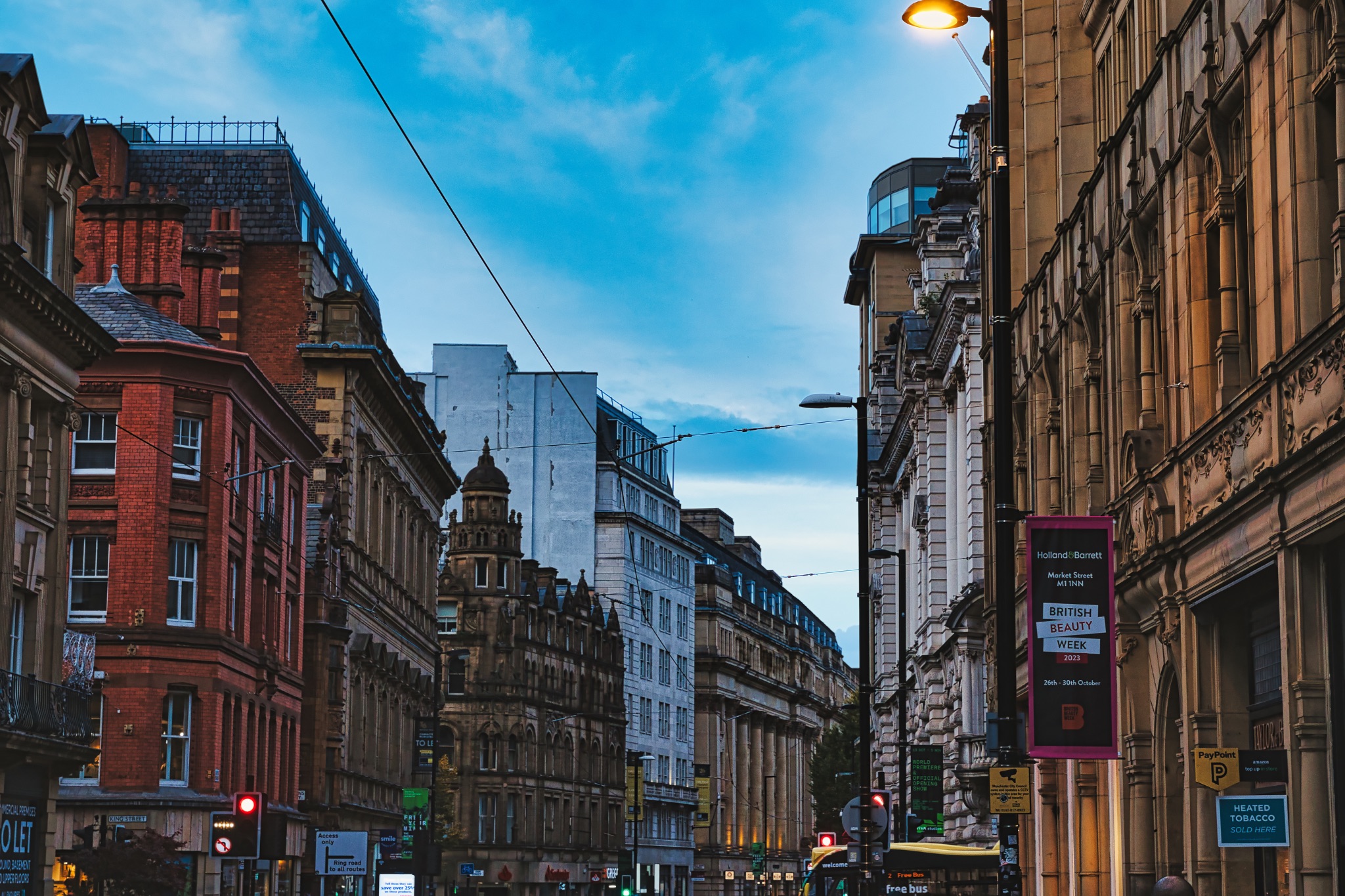 Dusk view of a bustling city street with historic architecture and glowing street lights under a blue sky in Manchester, England.