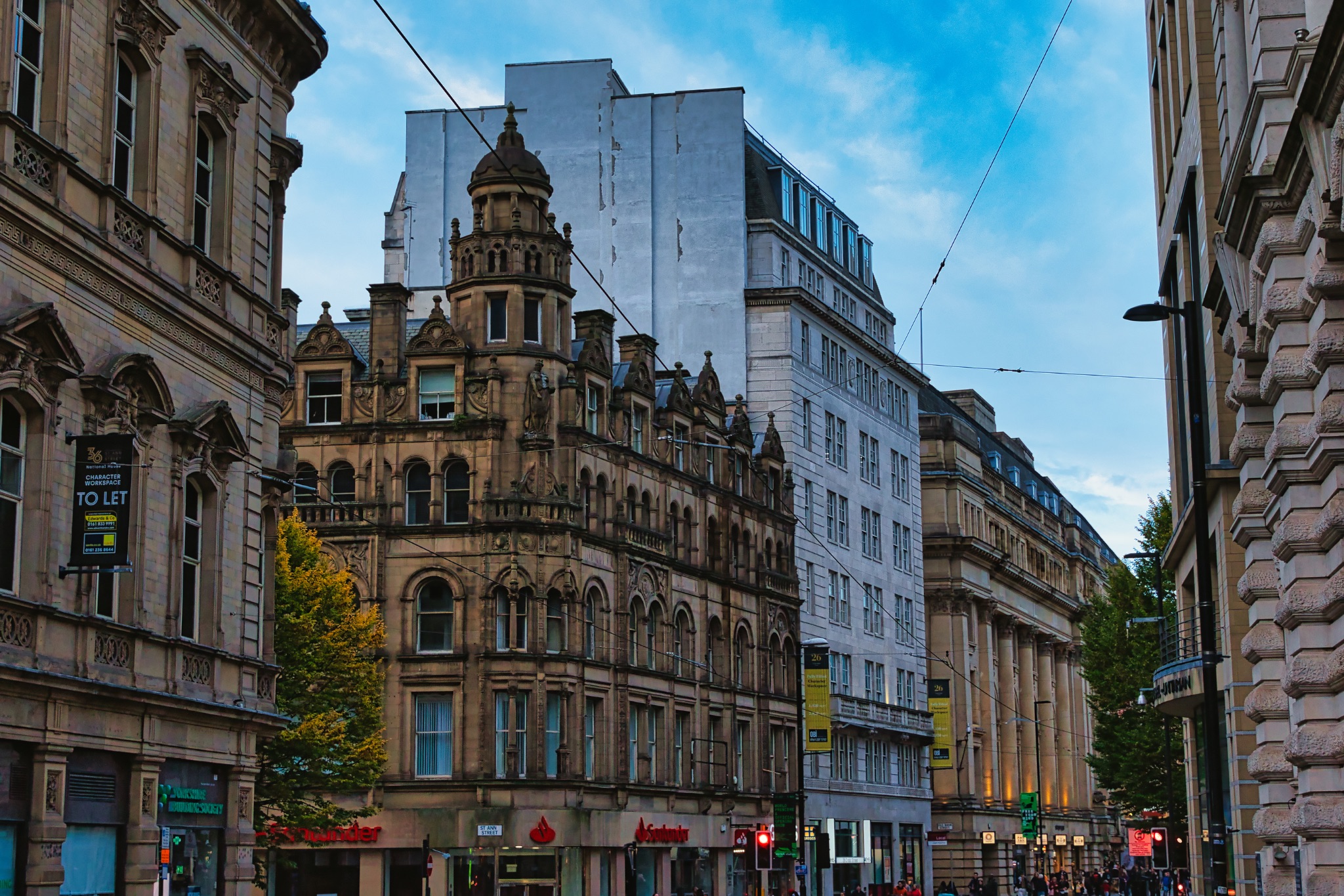 Dusk view of a bustling city street with historic architecture and glowing street lights under a blue sky in Manchester, England.
