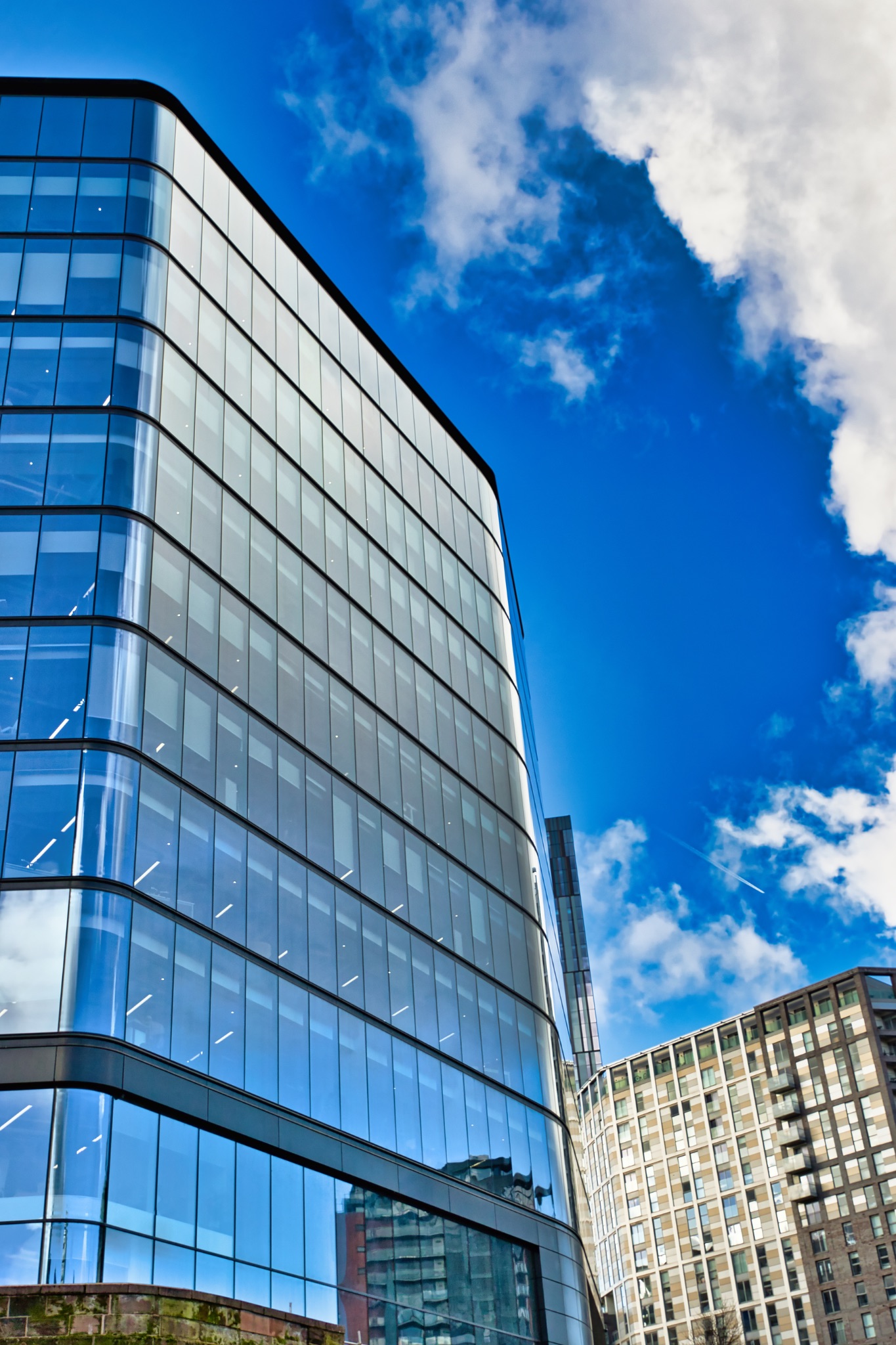Modern Glass Office Building and Adjacent Structures under a Blue Sky in Manchester, UK.