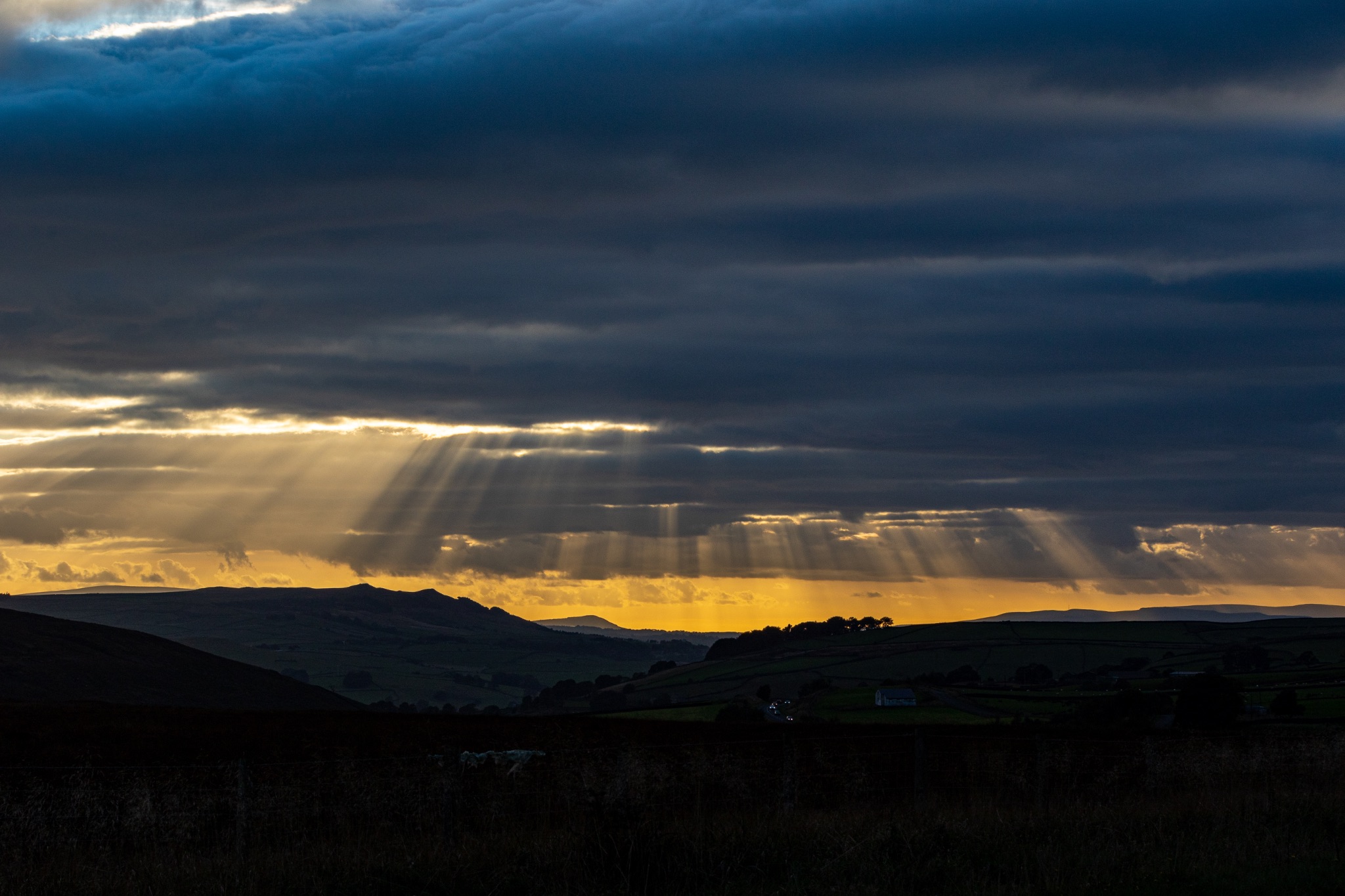 Dramatic sky with sunbeams piercing through clouds over a hilly landscape at dusk.