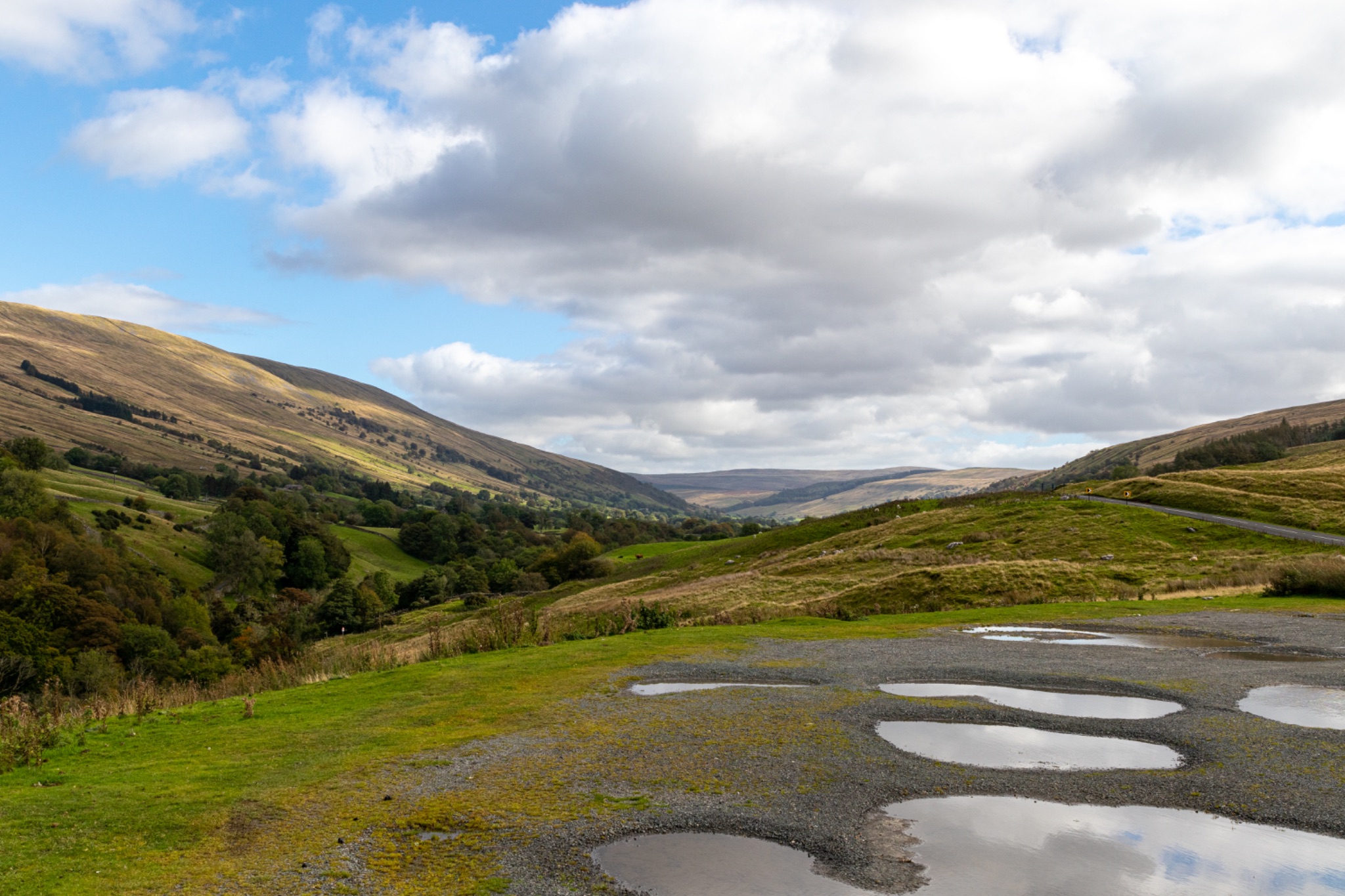 Scenic view of a lush valley with rolling hills under a cloudy sky, featuring a road with puddles after rain.