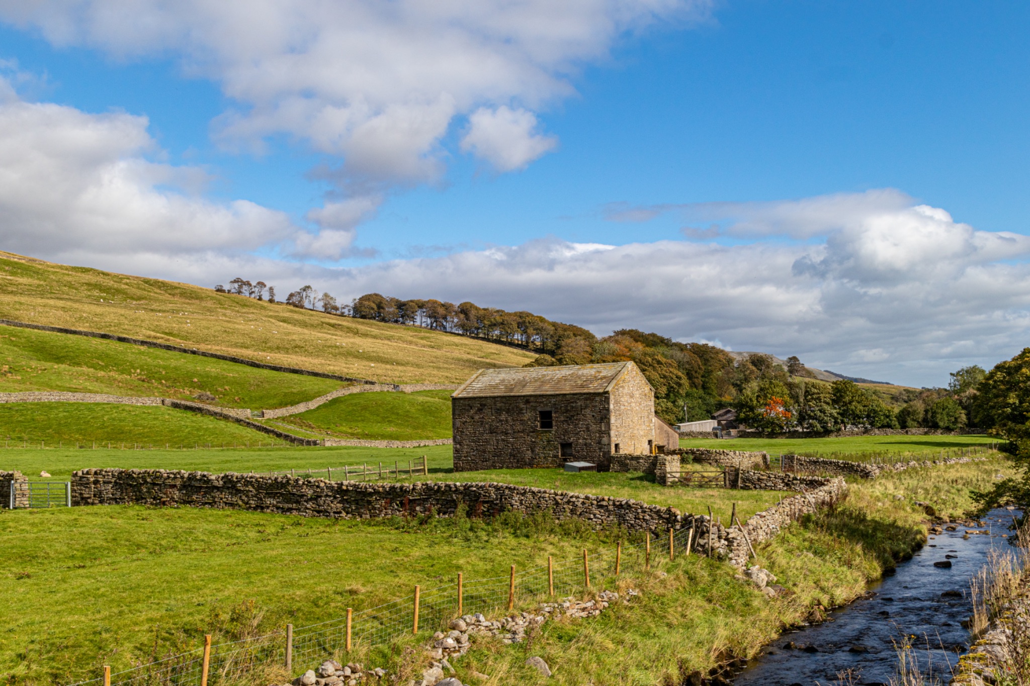 Idyllic countryside landscape with a stone barn, green fields, and a clear stream under a blue sky with fluffy clouds.