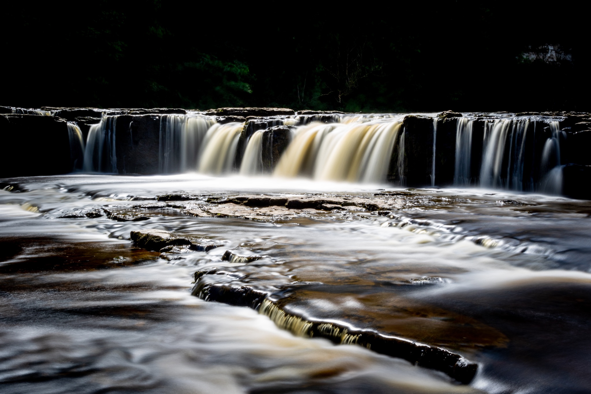 Silky water flows over a serene waterfall with dark rocks, captured in a long exposure photograph.