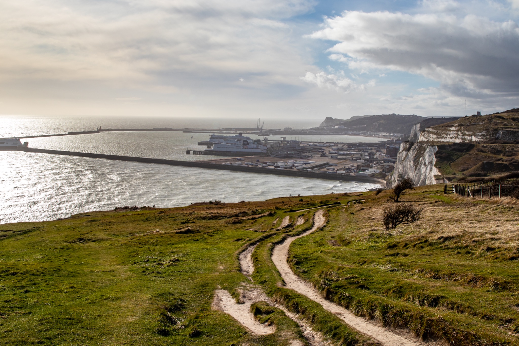 Scenic coastal landscape with winding footpath leading to a harbor under a cloudy sky.