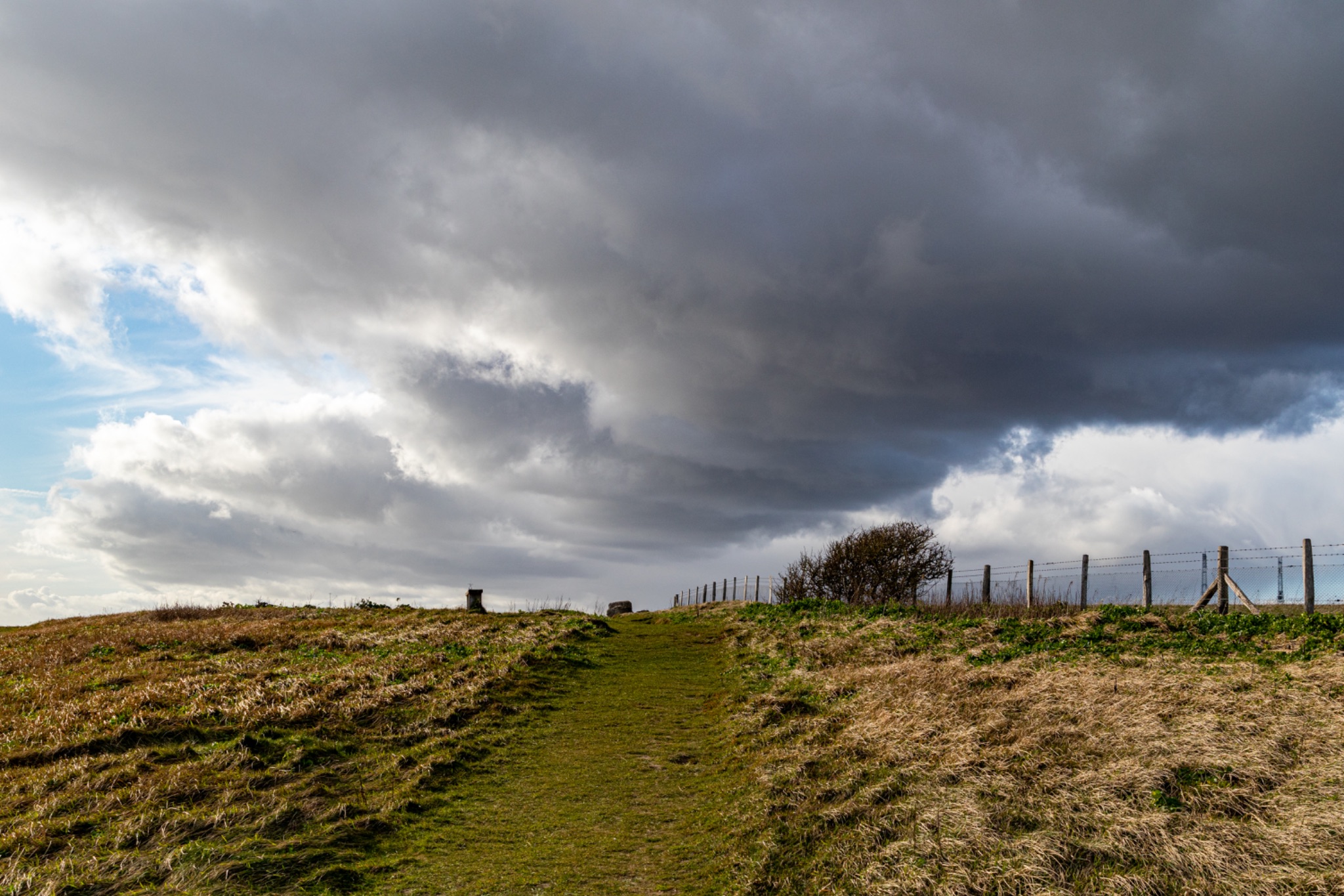Dramatic cloudscape over a rural landscape with a fence line and grassy path leading into the distance.