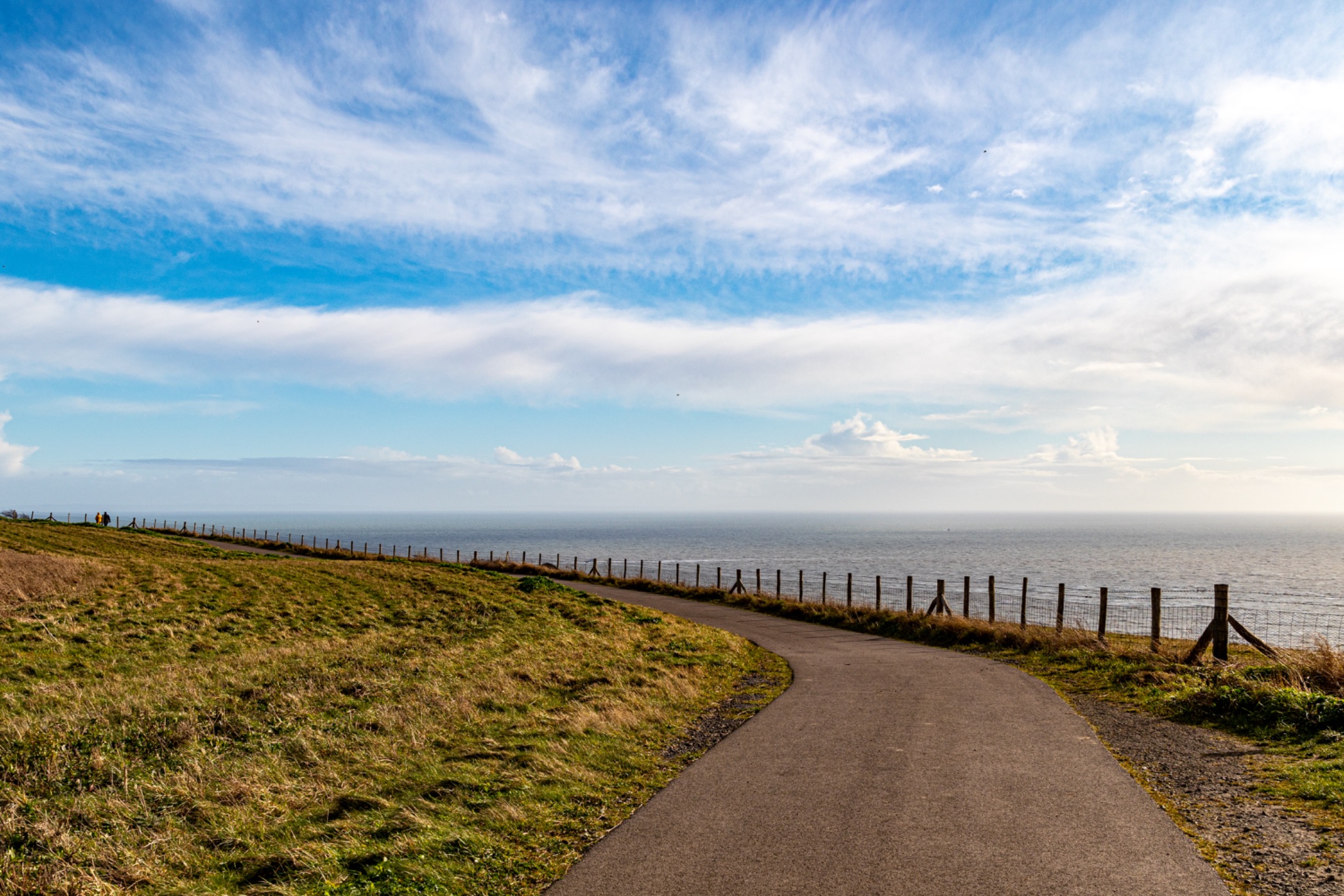 Scenic coastal pathway with wooden fence under a blue sky with fluffy clouds, leading towards the horizon over the ocean.