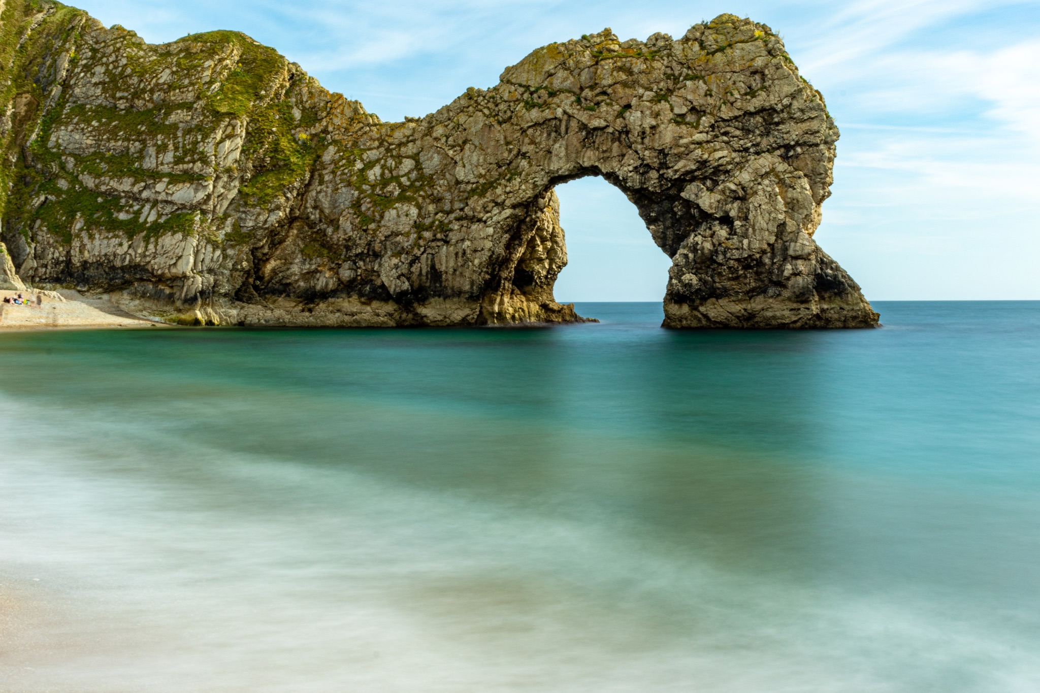 Serene seascape with a natural limestone arch on a sandy beach, clear turquoise waters under a blue sky at Durdle Door, England.
