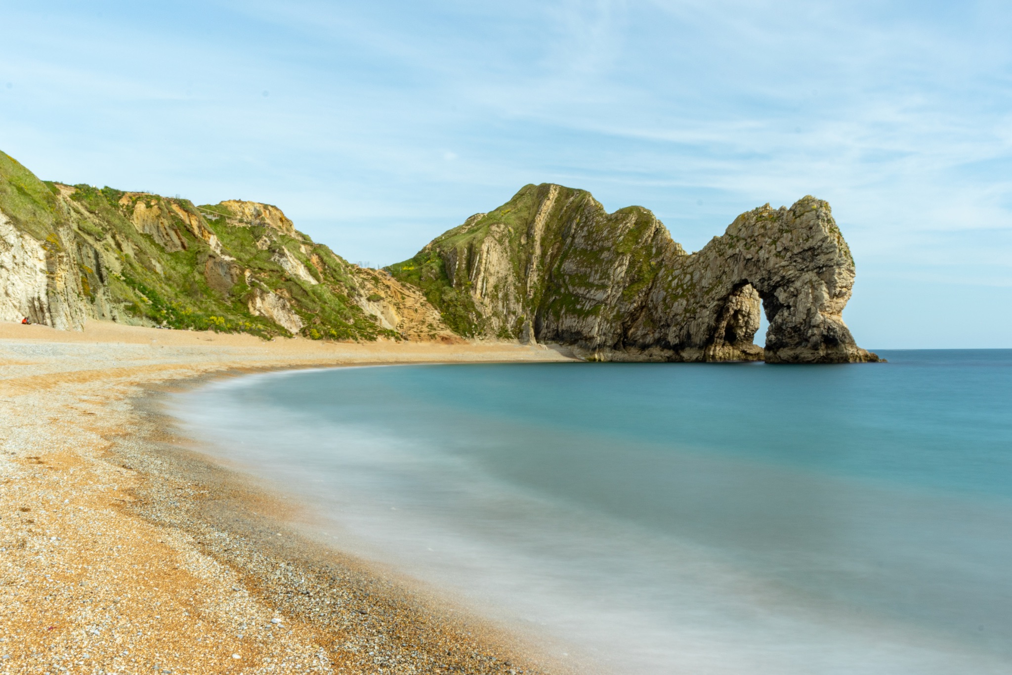 Serene beach with smooth water, pebbles, and rocky cliffs under a clear sky at Durdle Door, England.