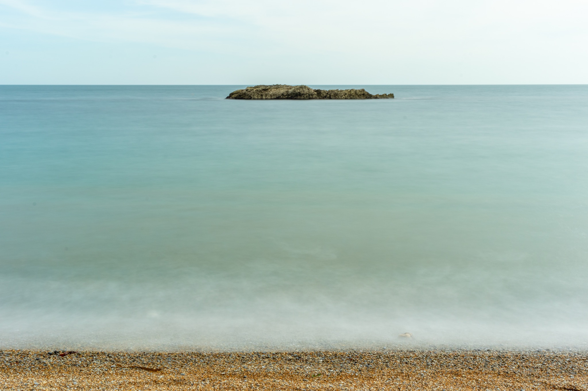 Serene seascape with a small island on the horizon, calm blue water, and a pebble beach in the foreground at Durdle Door, England.