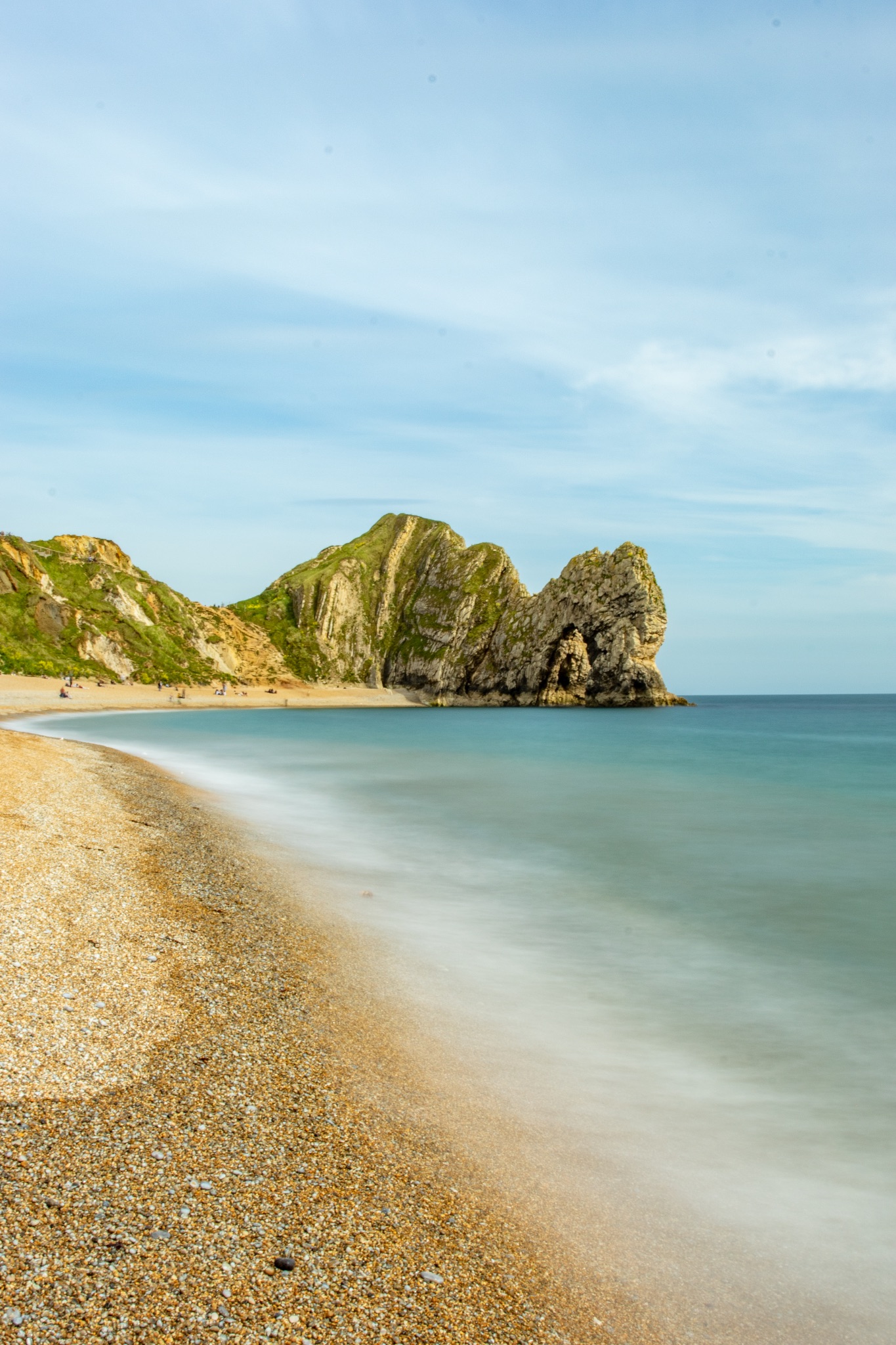 Serene beach with smooth water, pebbly shore, and rocky cliffs under a clear sky at Durdle Door, England.
