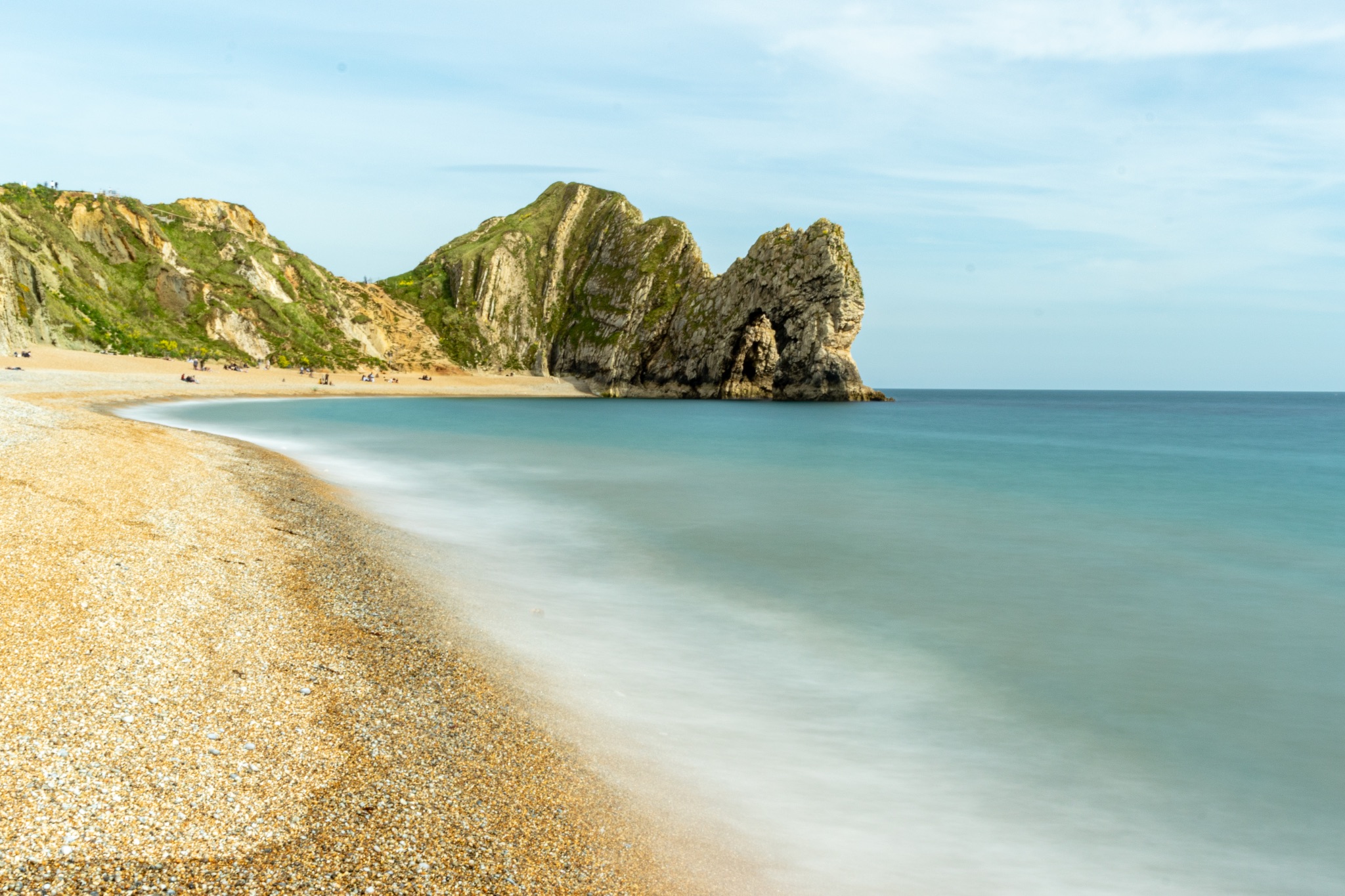 Serene beach with smooth water, pebbles, and rocky cliffs under a clear sky at Durdle Door, England.