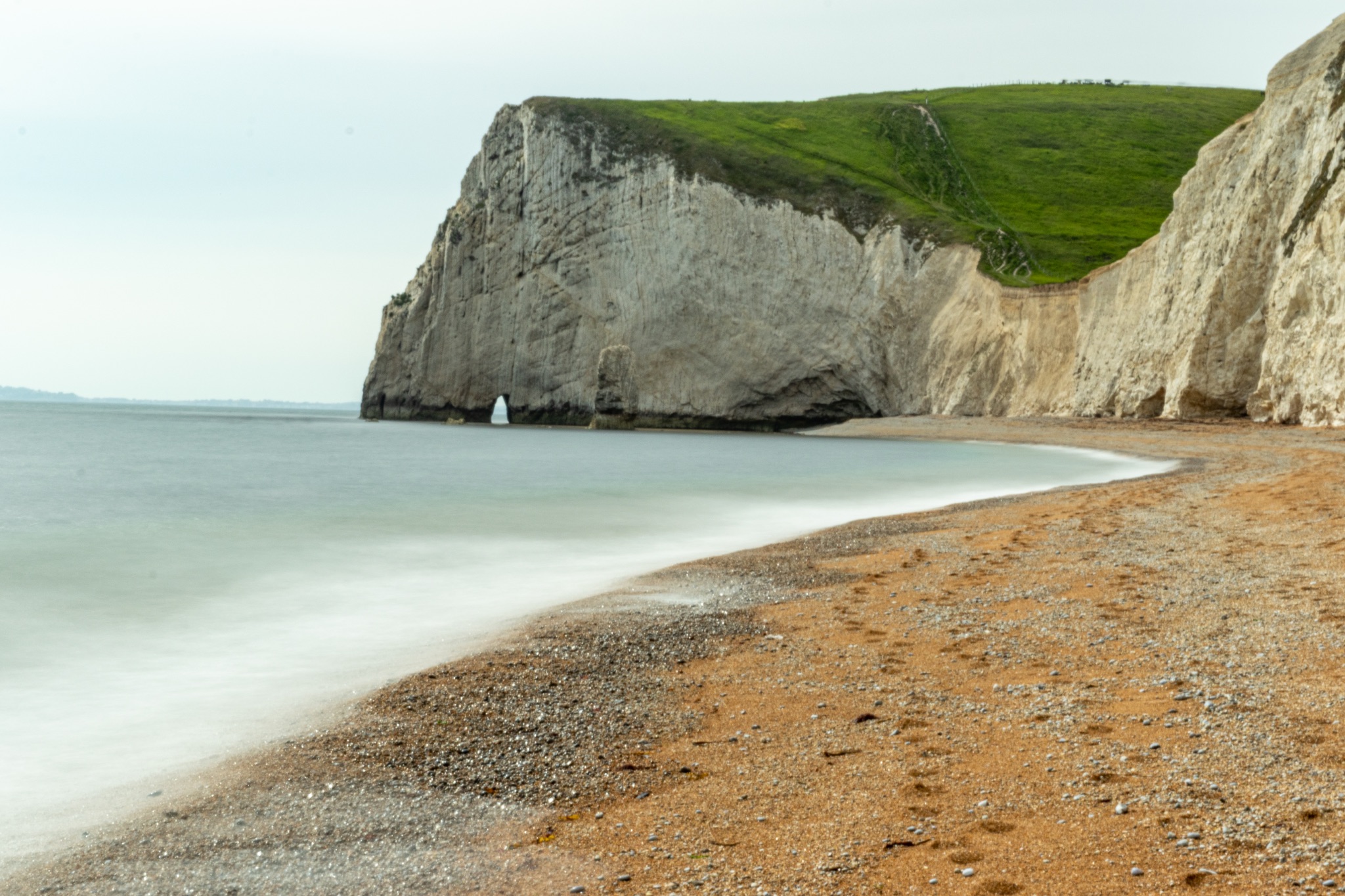 Serene beach with smooth waters, pebbles, and a towering cliff under a cloudy sky at Durdle Door, England.