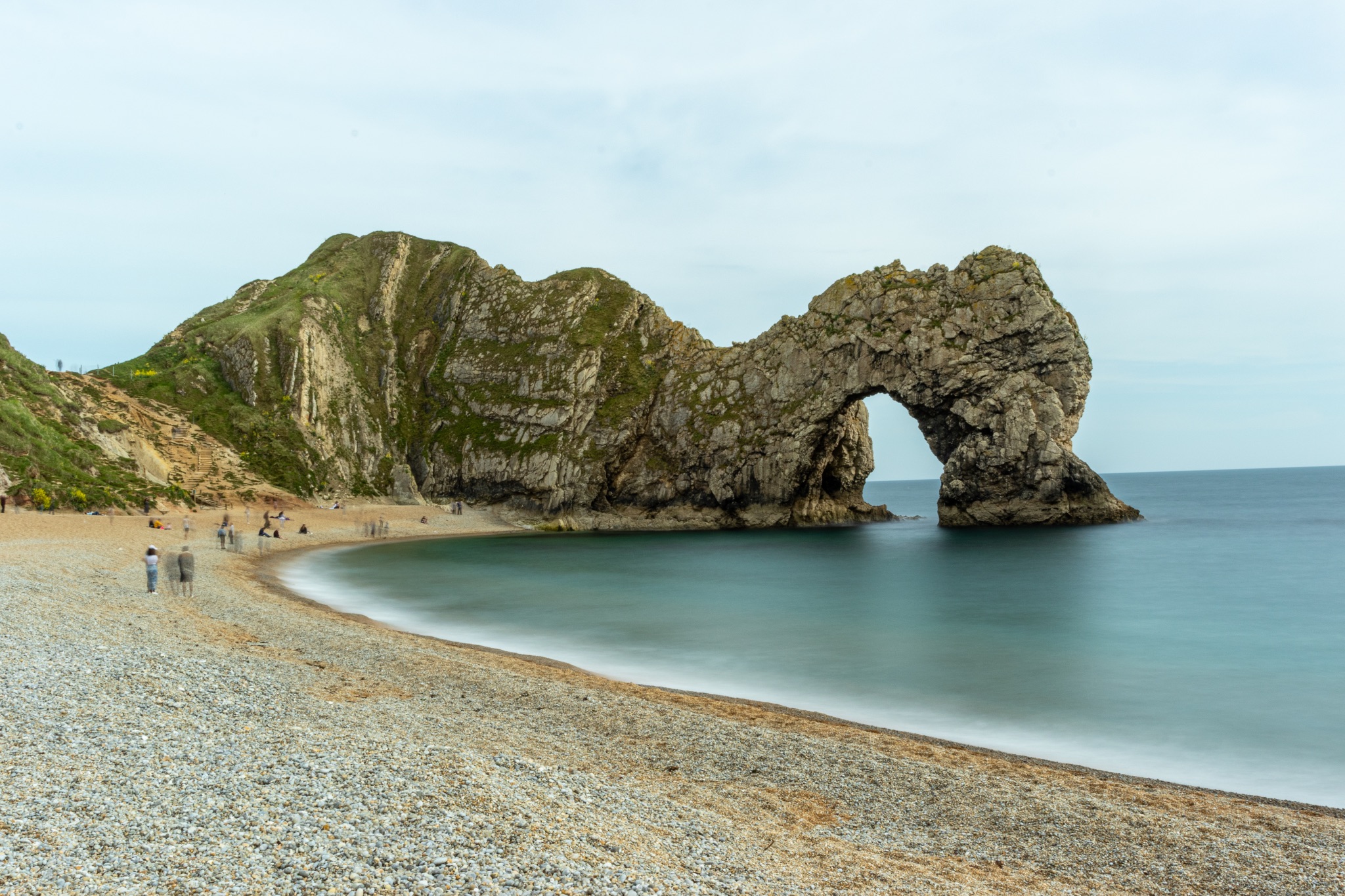 Scenic view of a natural arch on a pebble beach with calm sea and cloudy sky at Durdle Door, England.