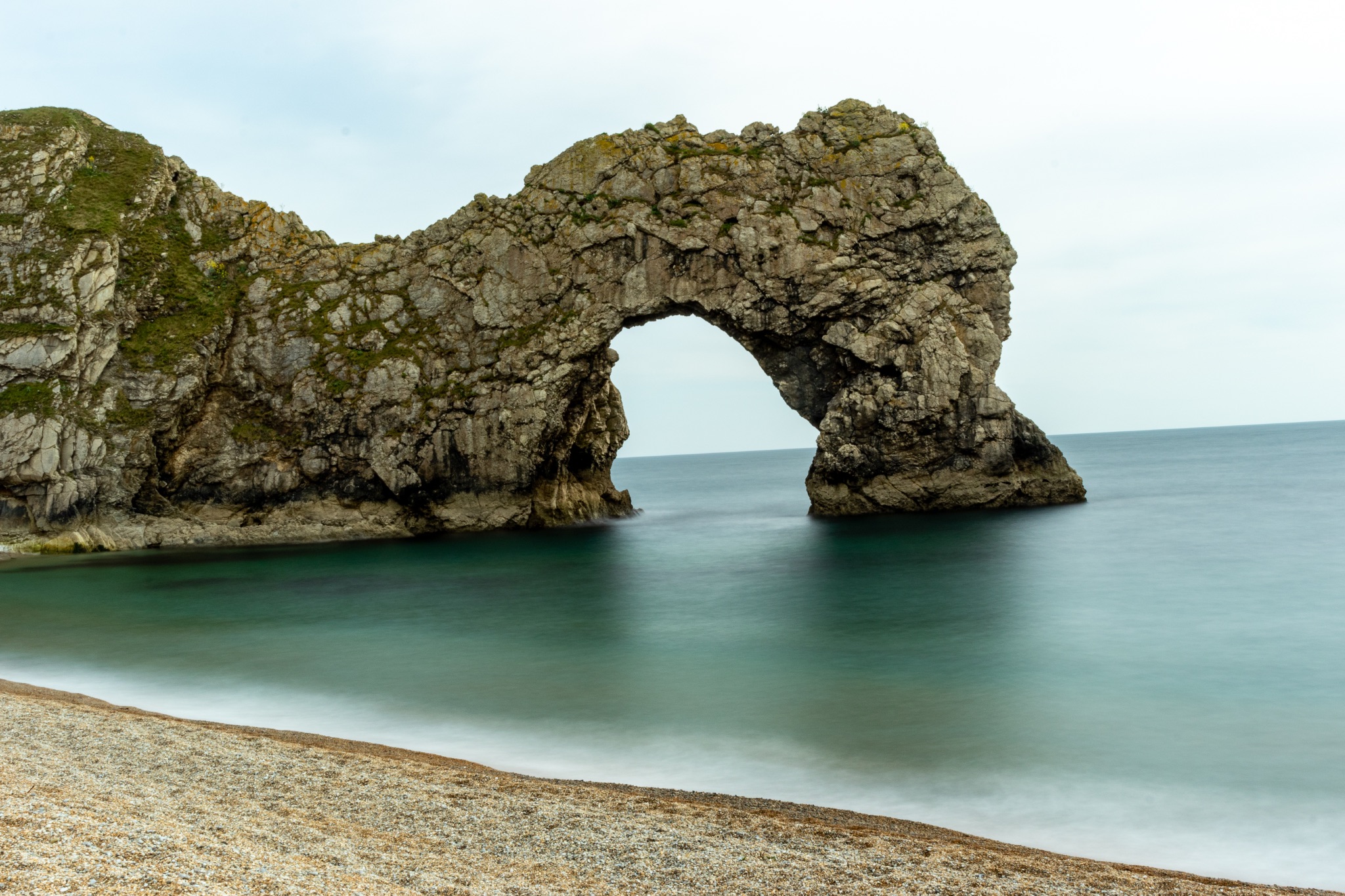Scenic view of a natural arch on a pebble beach with calm sea and cloudy sky at Durdle Door, England.