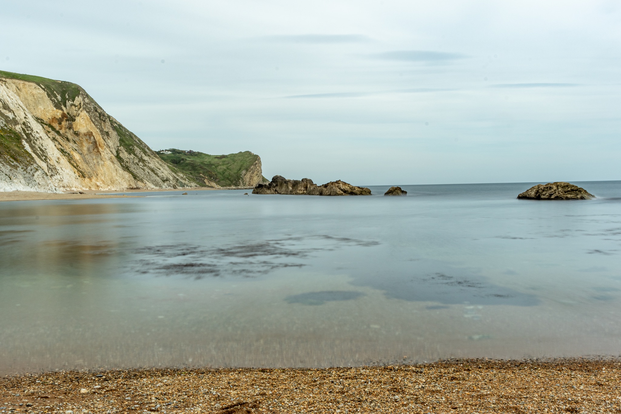 Tranquil beach scene with calm waters, sandy shore, and rocky cliffs under a soft cloudy sky at Durdle Door, England.