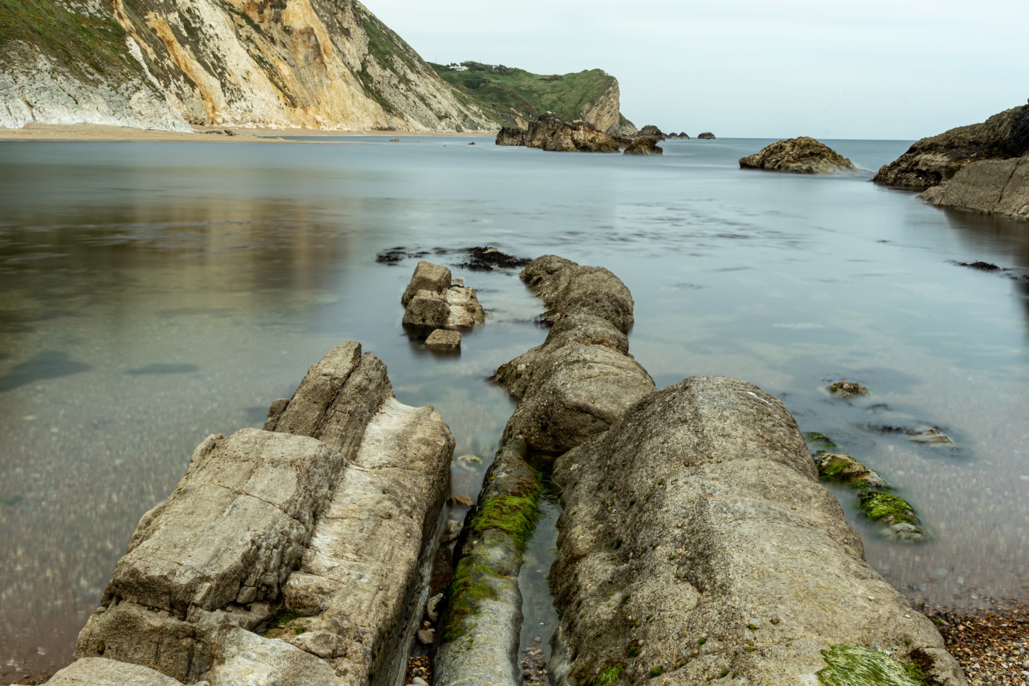 Serene coastal landscape with rocky outcrops leading into calm waters, surrounded by cliffs under a soft sky at Durdle Door, England.