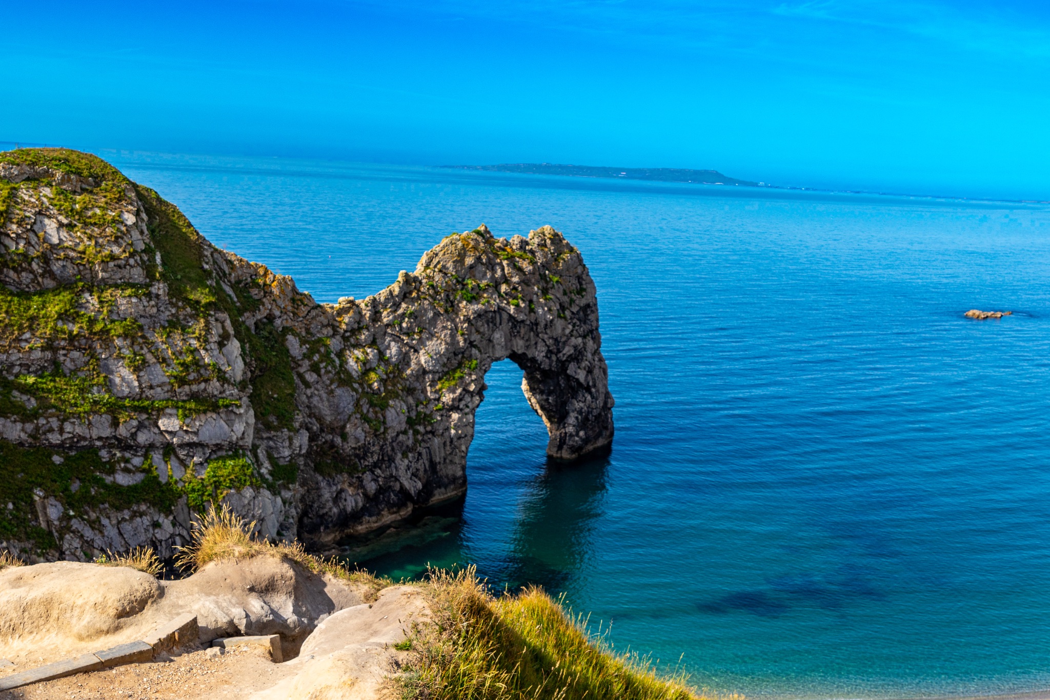 Scenic view of a natural arch on a coastline with a sandy beach and clear blue sky at Durdle Door, England.