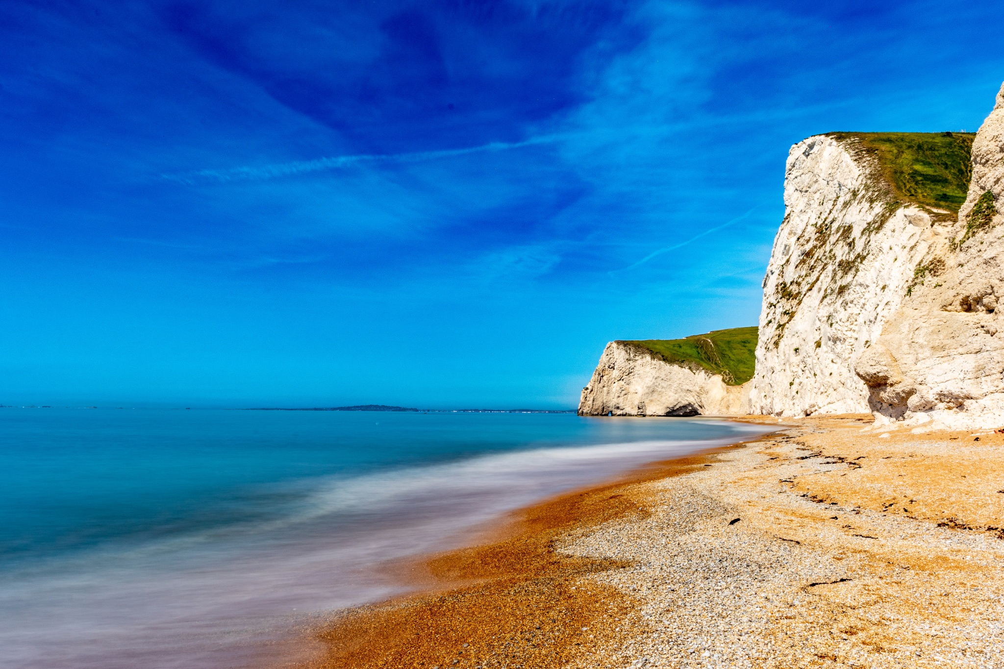 Serene beach with smooth water effect, blue skies, and towering cliffs on a sunny day at Durdle Door, England.