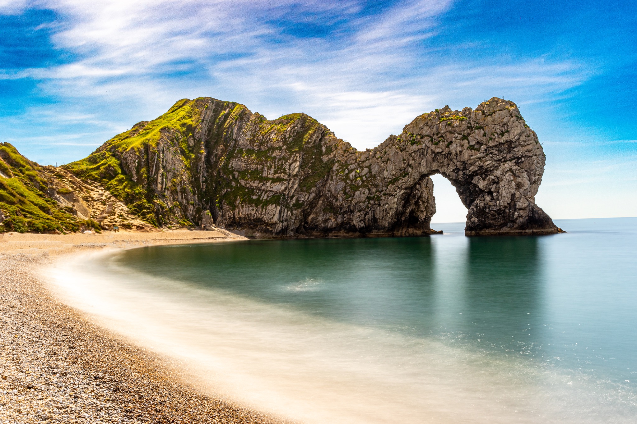Scenic view of a natural arch on a coastline with a sandy beach and clear blue sky at Durdle Door, England.