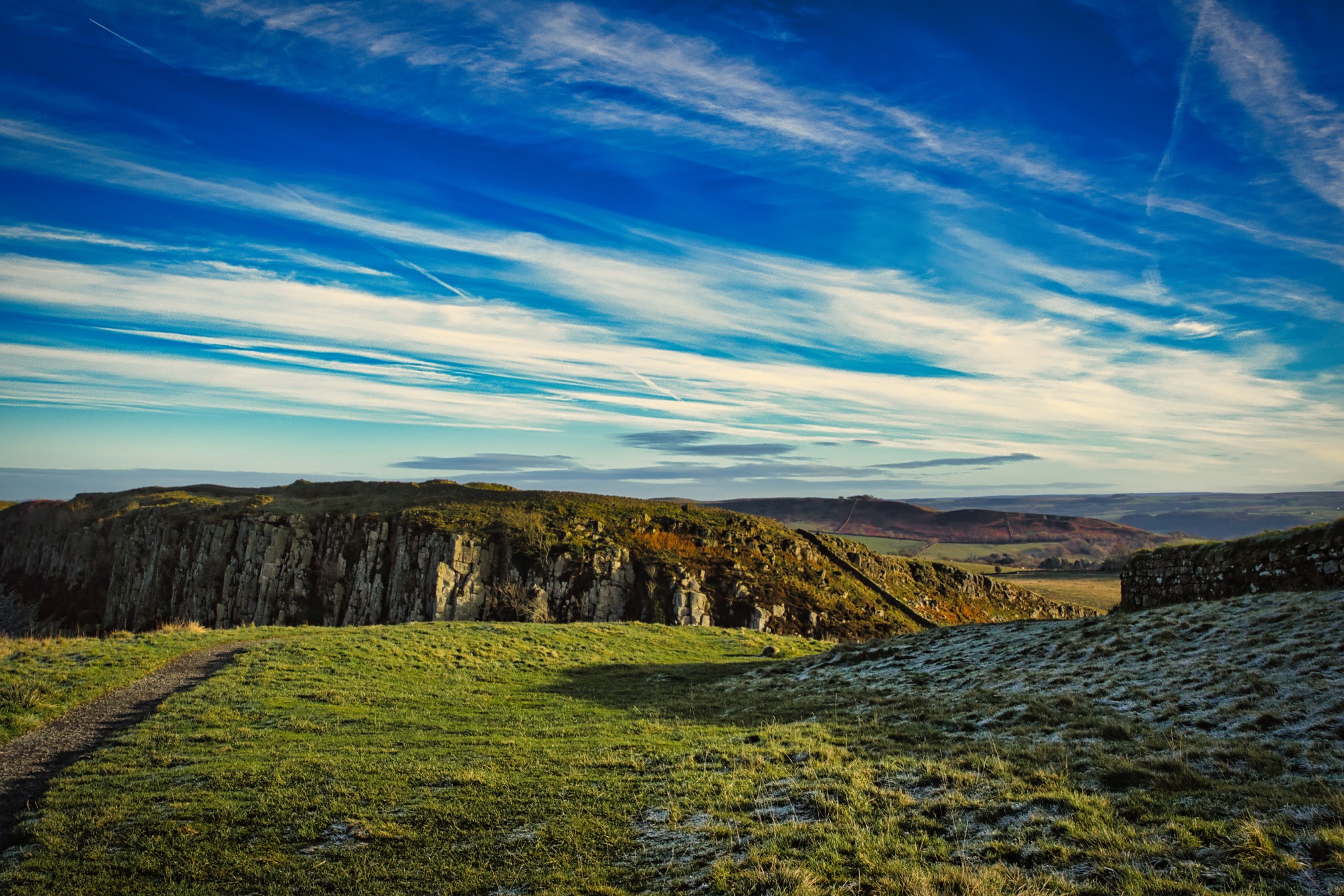 Breathtaking landscape with rocky cliffs under a vibrant blue sky with wispy clouds, green grass in the foreground at Sycamore Gap, Northumberland, UK.