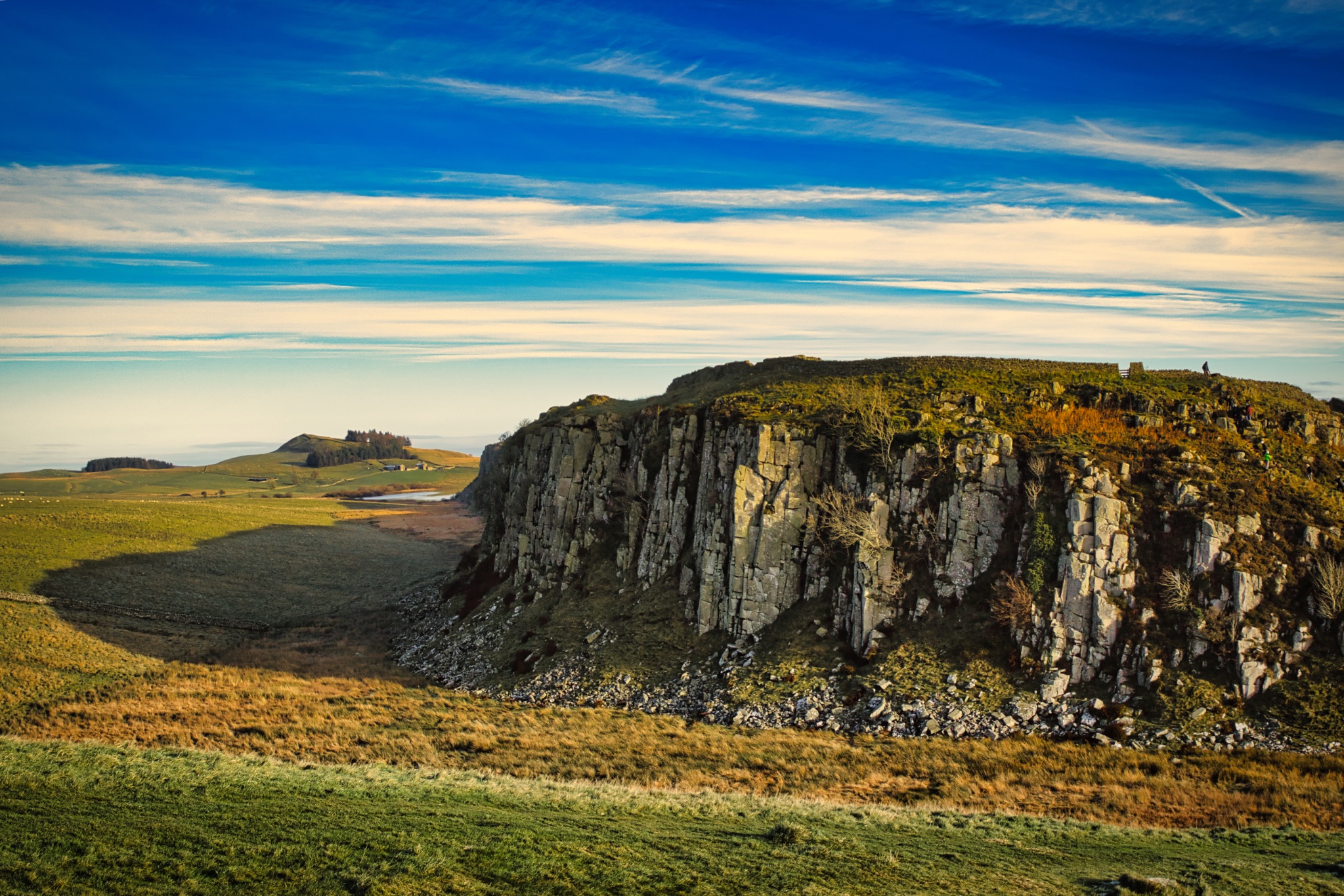 Scenic view of a rugged cliff against a blue sky with wispy clouds, highlighting nature's grandeur at Sycamore Gap, Northumberland, UK.