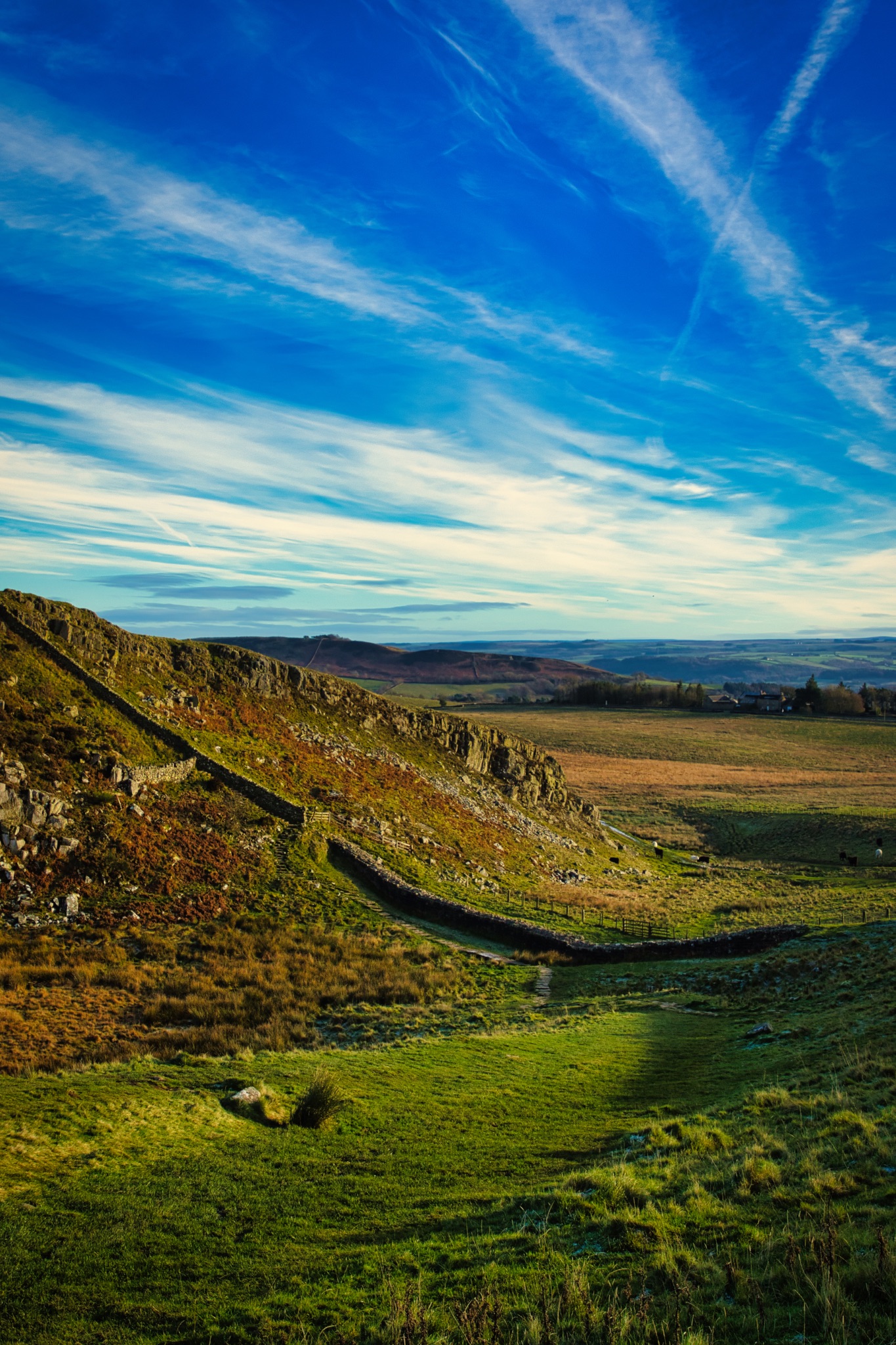 Breathtaking landscape with vibrant blue skies, wispy clouds, and a scenic rocky hillside leading to a lush green valley at Sycamore Gap, Northumberland, UK.