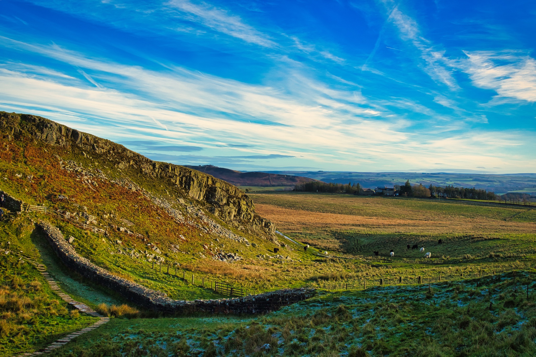Scenic landscape with vibrant blue skies, wispy clouds, and a rocky hillside overlooking a lush green valley at Sycamore Gap, Northumberland, UK.