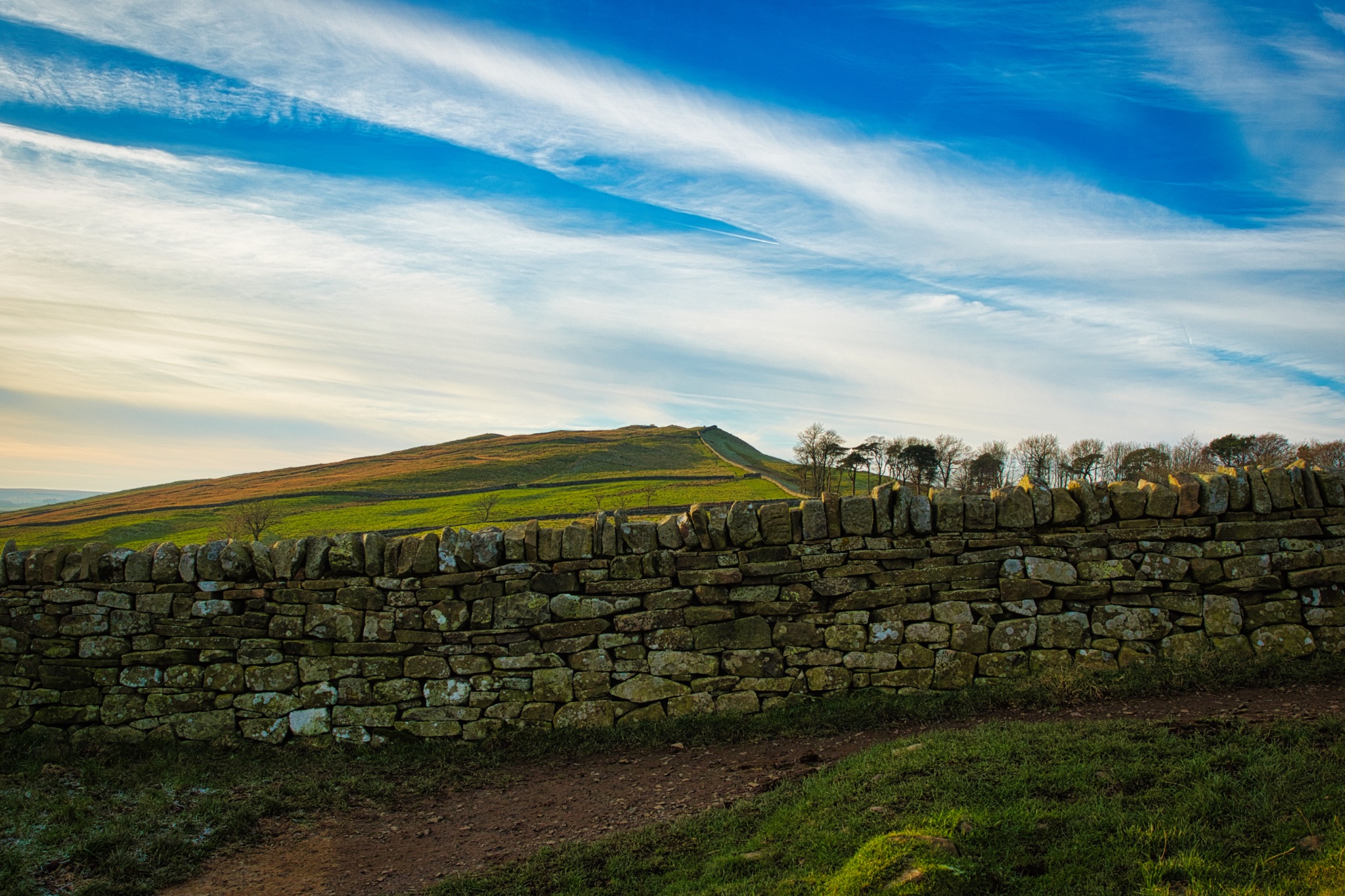 Scenic view of a dry stone wall leading towards a hill under a dramatic sky at sunset.