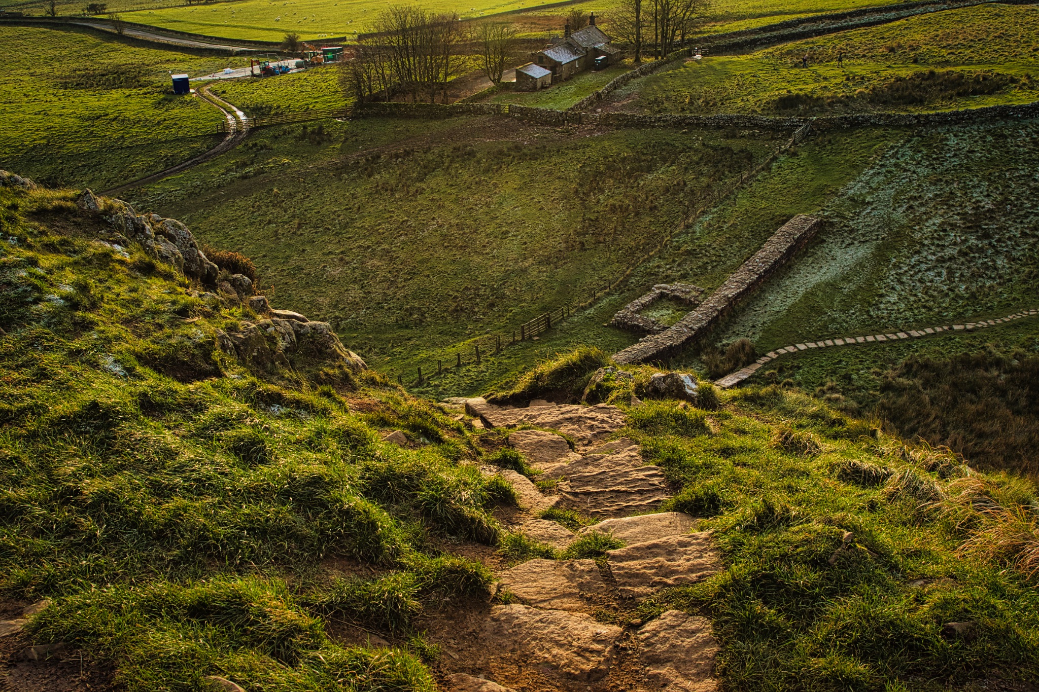 Rustic countryside path leading through rolling green hills with a small village and winding road in the distance at sunset at Sycamore Gap, Northumberland, UK.